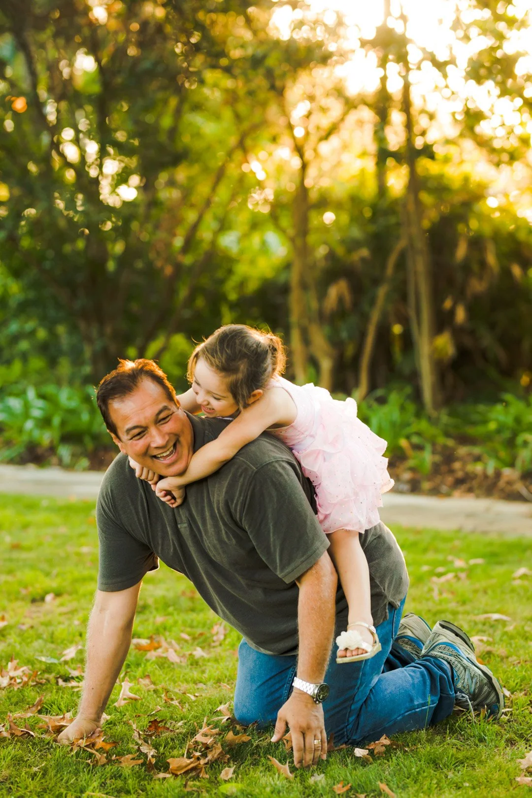 A man kneeling on grass, smiling, as a young girl in a pink dress playfully climbs on his back outdoors during sunset.