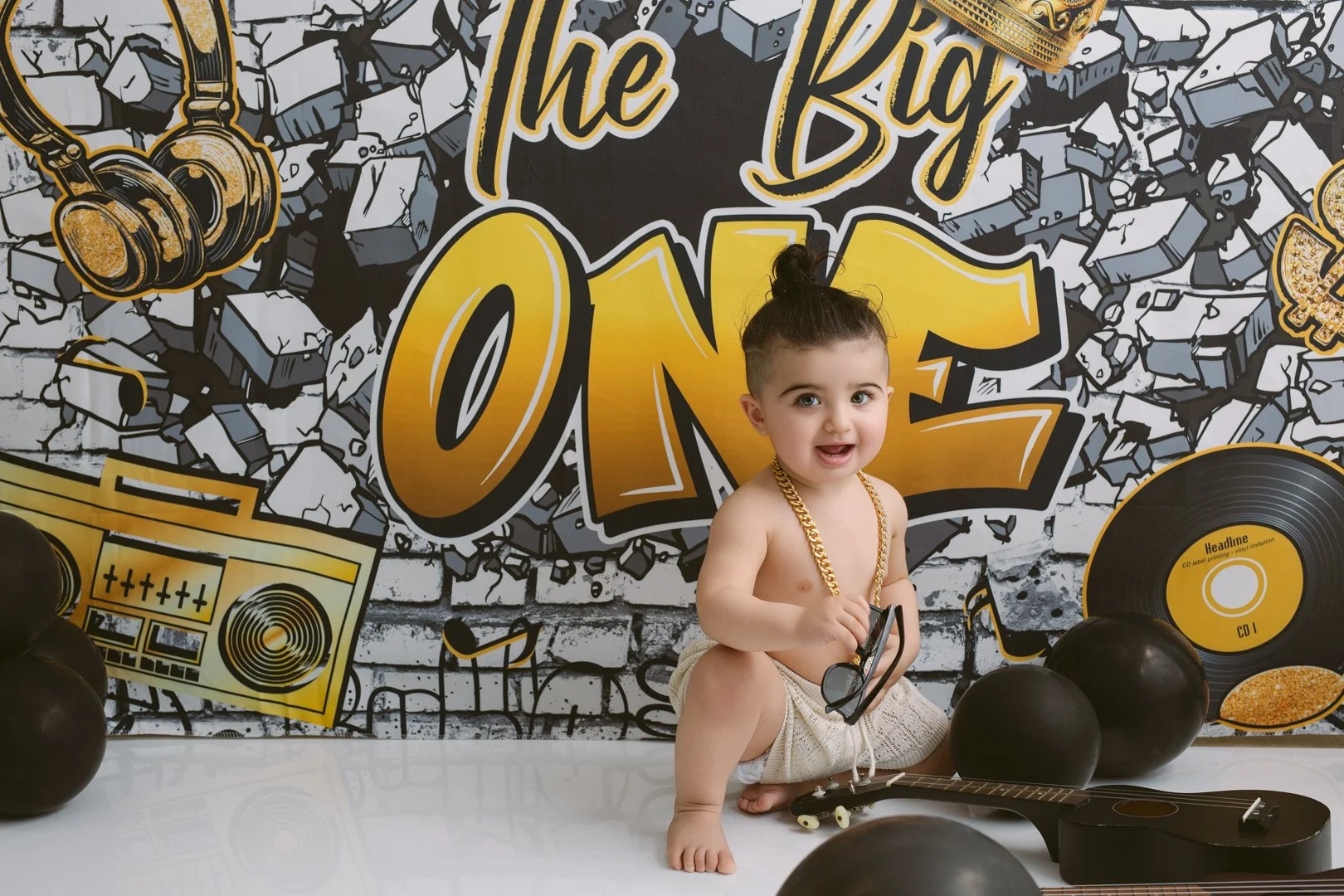 Young child sitting on the floor at a party, wearing a gold chain and holding sunglasses, with a guitar in front. The background features a wall with graffiti-style artwork and large yellow text reading 'The Big ONE,' surrounded by balloons and recor