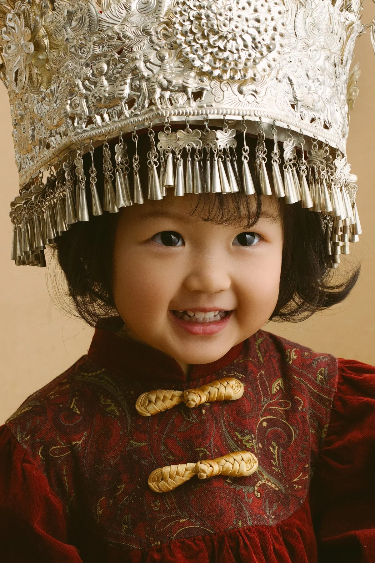 A young girl smiling, wearing a red traditional outfit with gold decorative clasps, and a silver ornate hat with dangling elements.