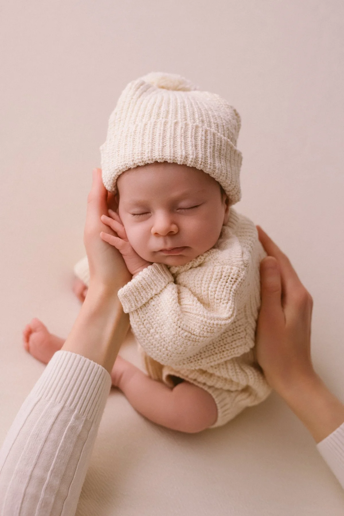 A newborn baby sleeping peacefully with a soft cream-colored knit hat and sweater, gently held by adult hands.