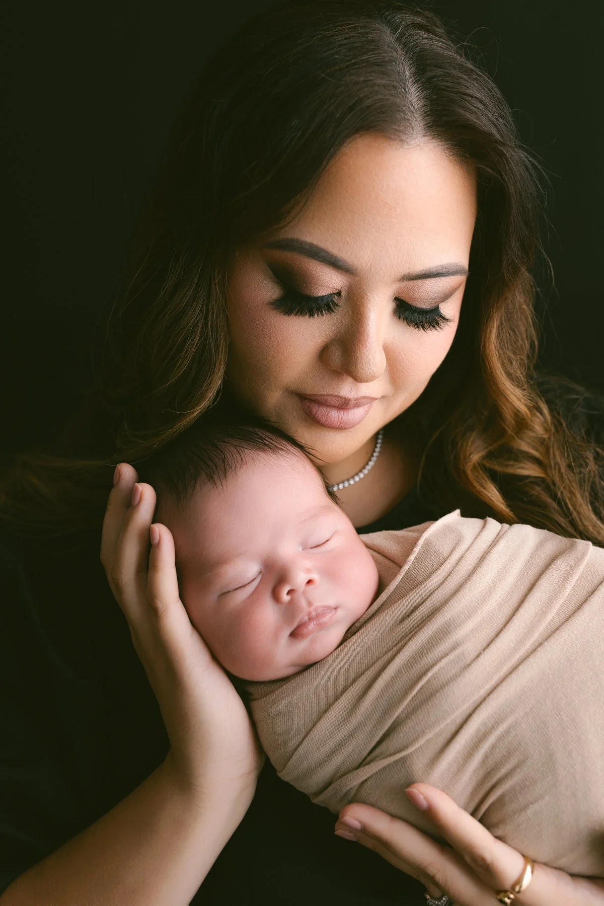 A woman with long brown hair and makeup holding a sleeping baby, wrapped in a beige blanket, against a dark background.