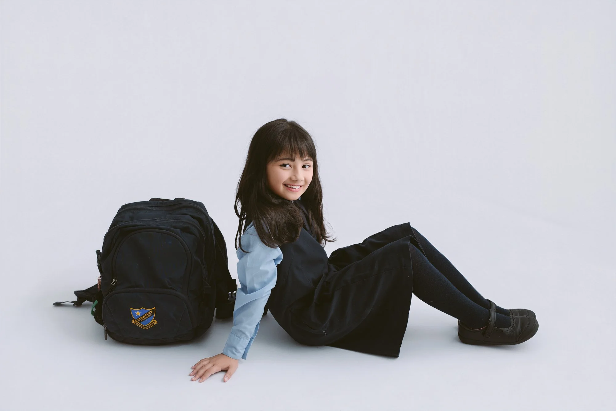 A young girl with dark hair and a light blue shirt under a navy dress, smiling and sitting on the floor against a plain white background, with a black backpack beside her.