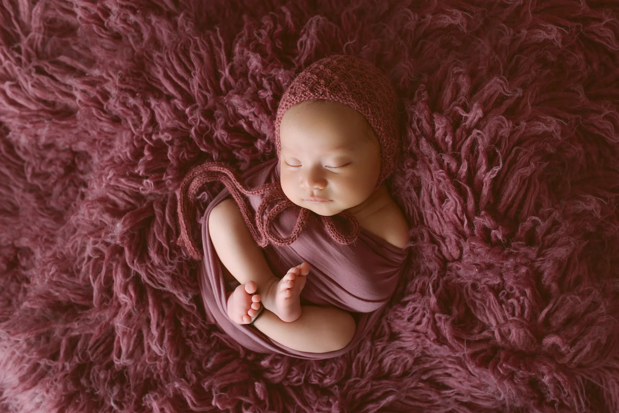 A newborn baby sleeping on a fluffy, mauve-colored blanket, wearing a matching crochet hat and wrap.
