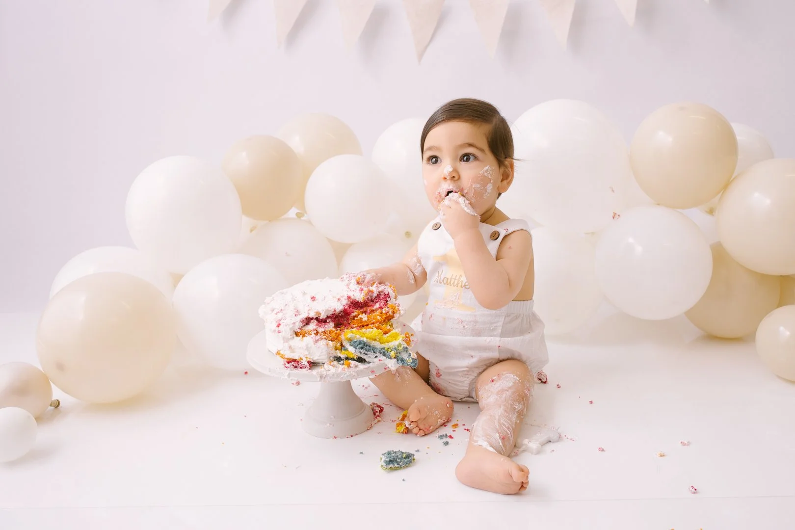 A young child with cake and white frosting on face and body sitting on the floor surrounded by white balloons, with a cake on a stand and colorful cake inside.