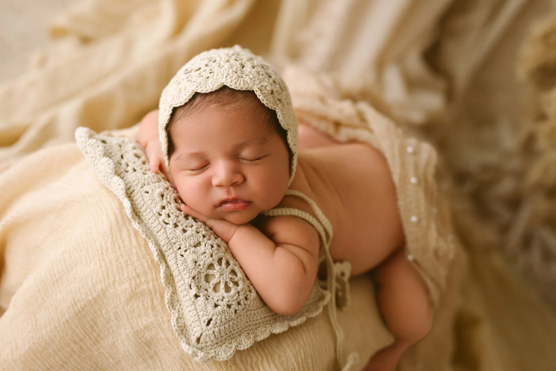 A sleeping baby wearing a crocheted hat and matching outfit, lying on a soft, beige blanket.