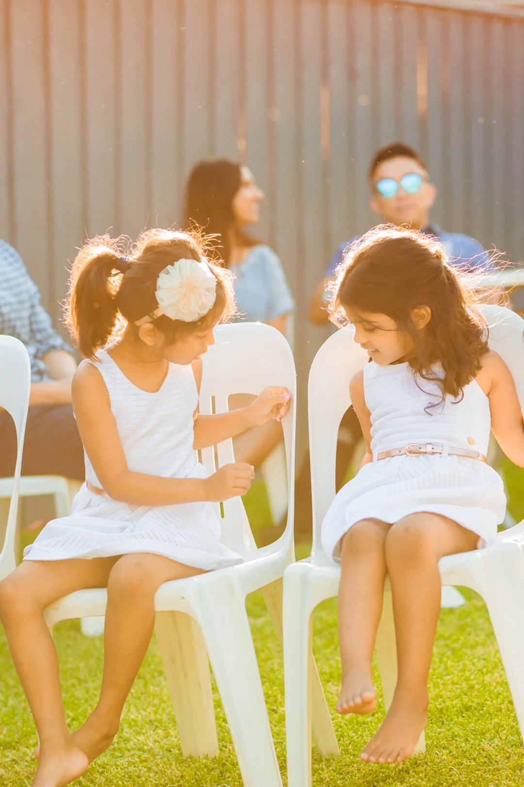 Two young girls in white dresses sitting barefoot on white plastic chairs on a grassy lawn, engaged in a conversation, with adults seated behind them, blurred in the background, during a sunny outdoor event.