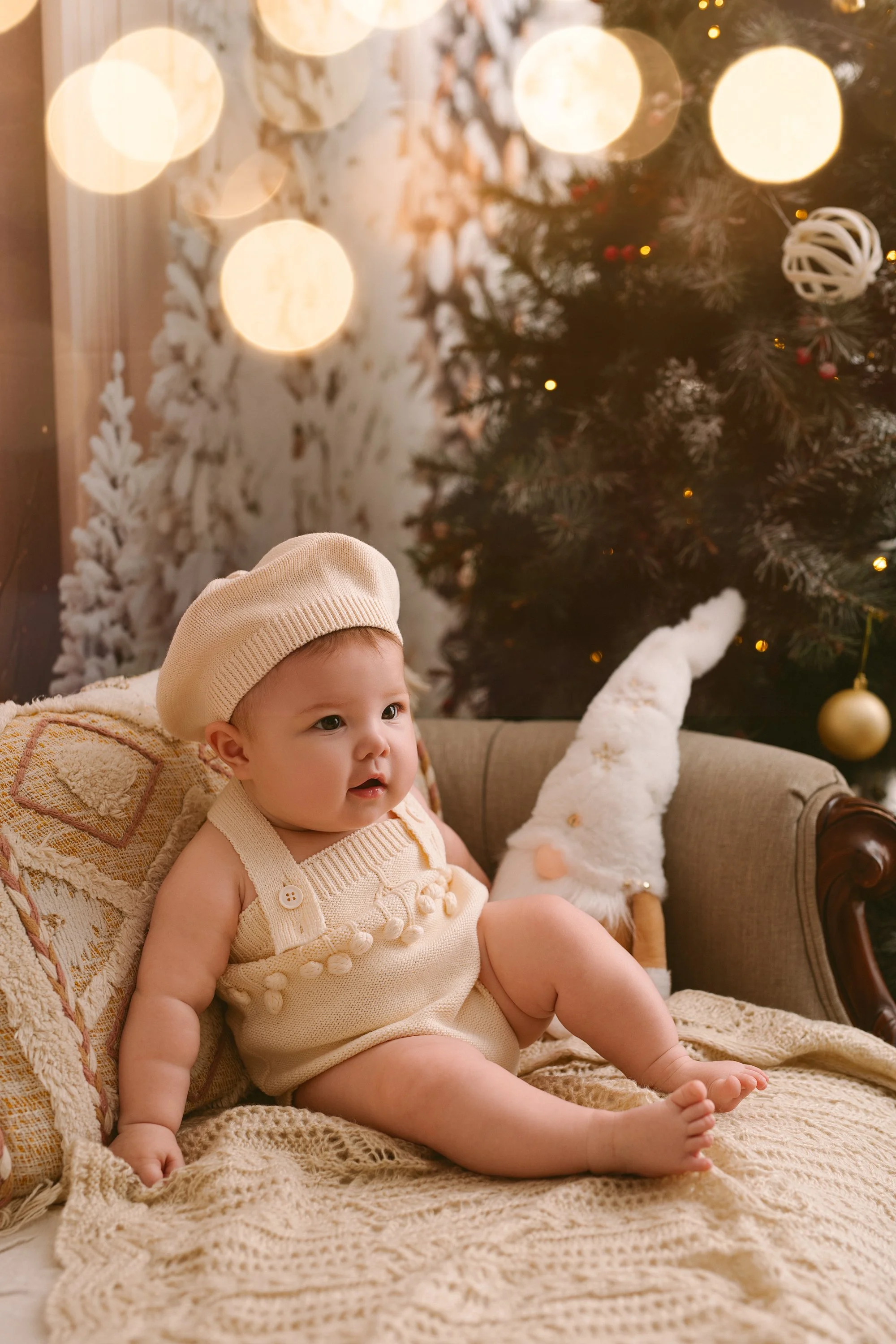 A baby sitting on a beige knitting blanket on a sofa, wearing a cream-colored knitted hat and romper, with Christmas decorations including a Christmas tree with ornaments and lights, and a plush gnome figure in the background.