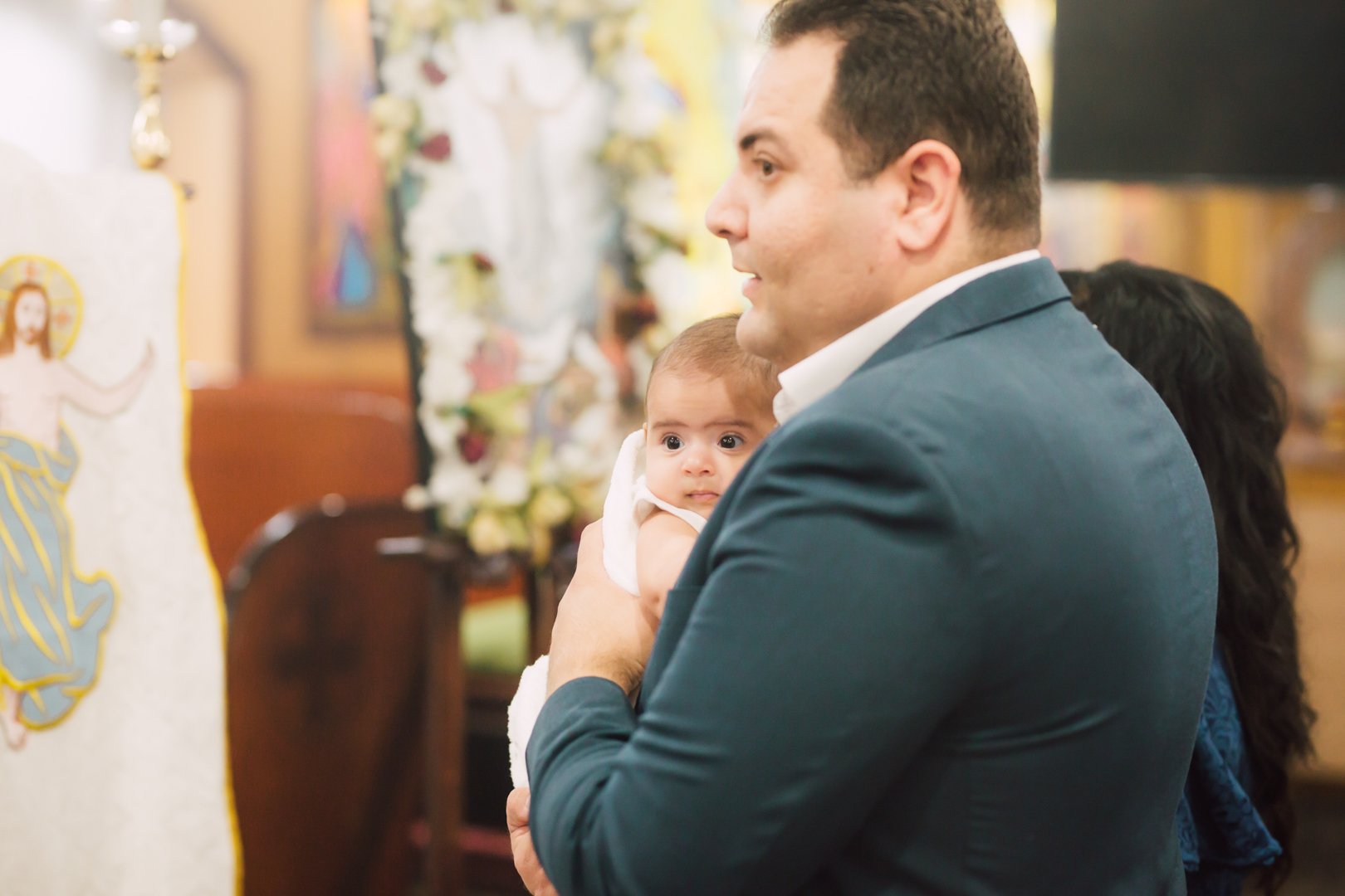 A man in a suit holding a baby girl during a religious ceremony, with religious artwork visible in the background.