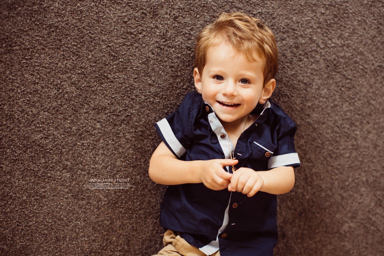 A young boy with short light brown hair, wearing a navy blue short-sleeved shirt with white accents and tan pants, smiling while lying on a brown carpet.