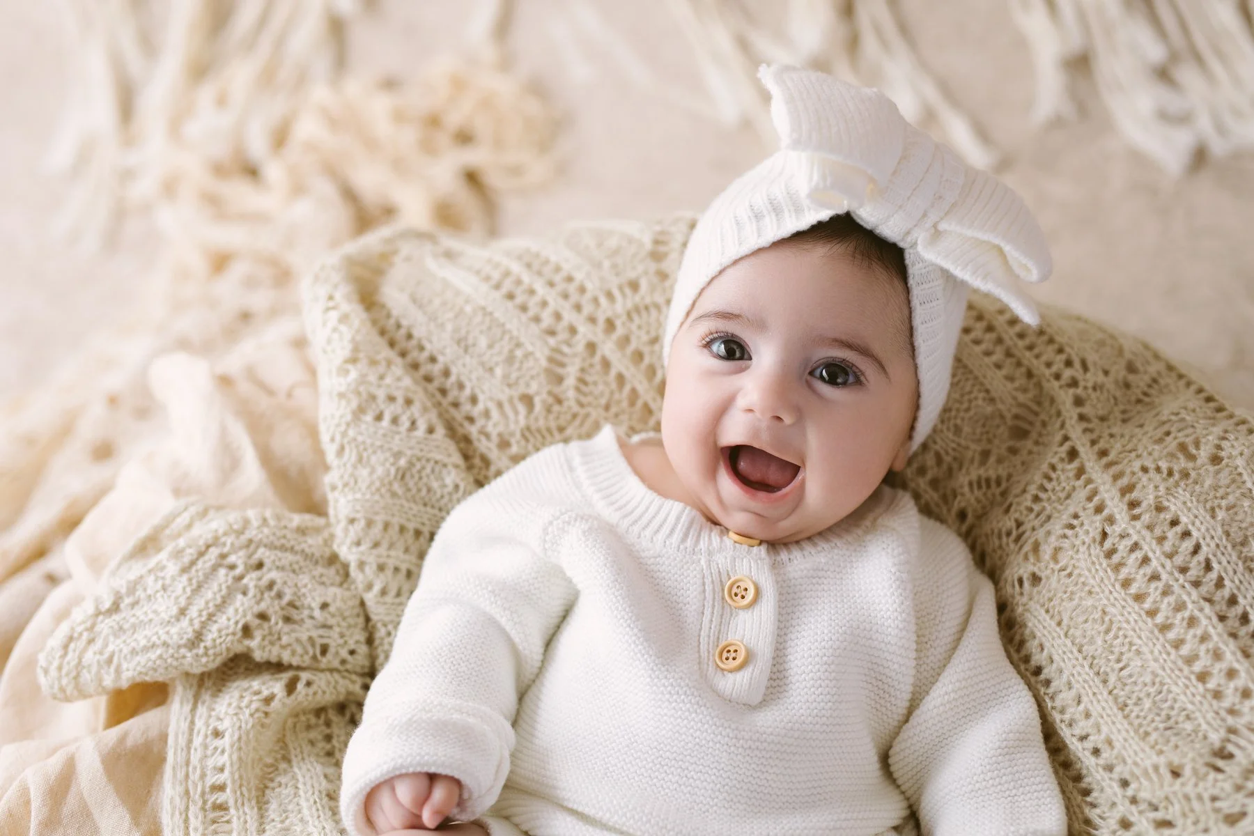 A smiling baby wearing a white knit sweater and a white knit hat with a bow, lying on a cream-colored, textured blanket.