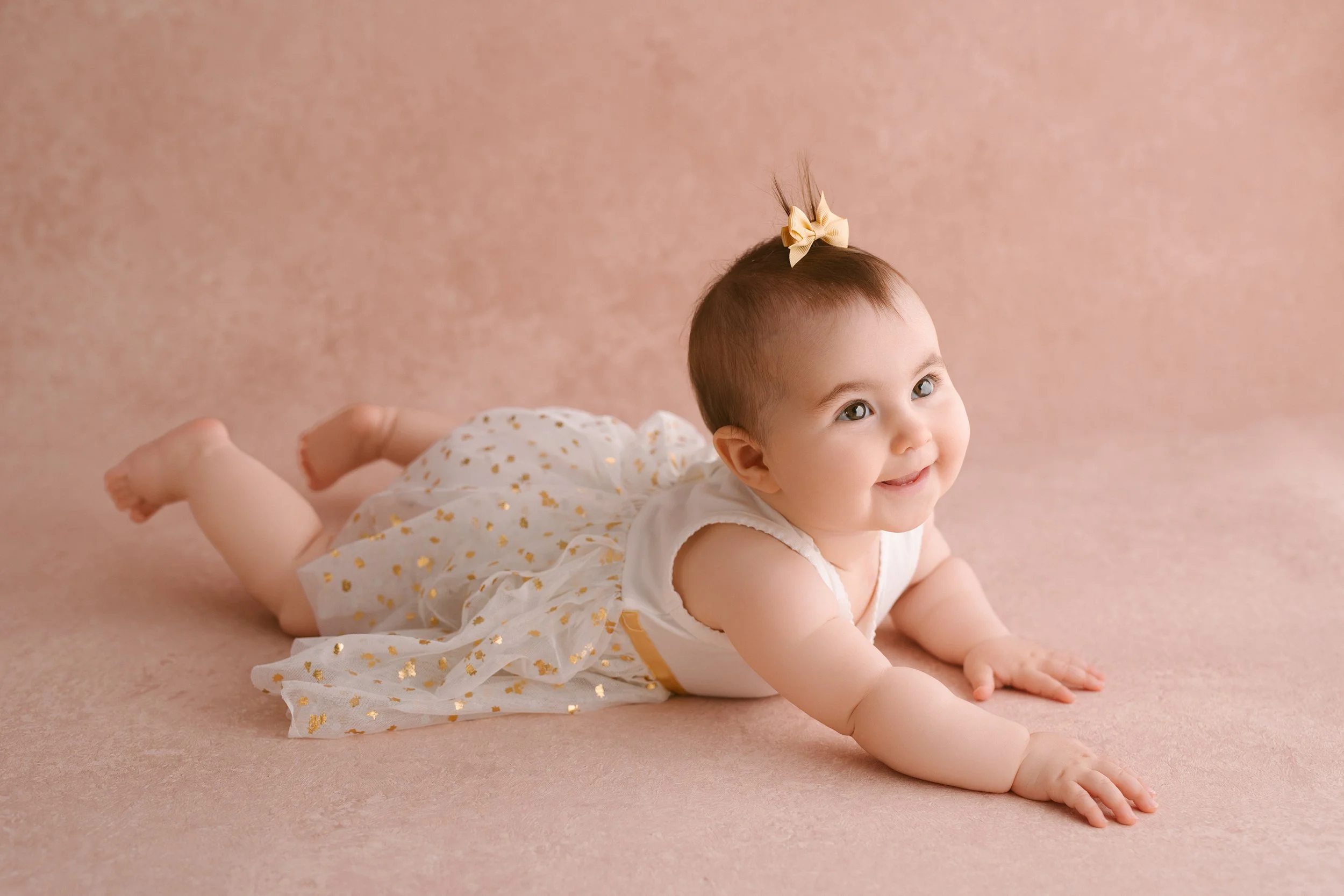 A smiling baby girl with a yellow bow in her hair, lying on her stomach on a pink textured background, wearing a white dress with gold accents.