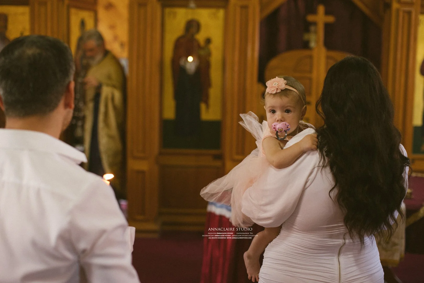 A young girl with a pacifier in her mouth being held by a woman inside a church with religious icons in the background.