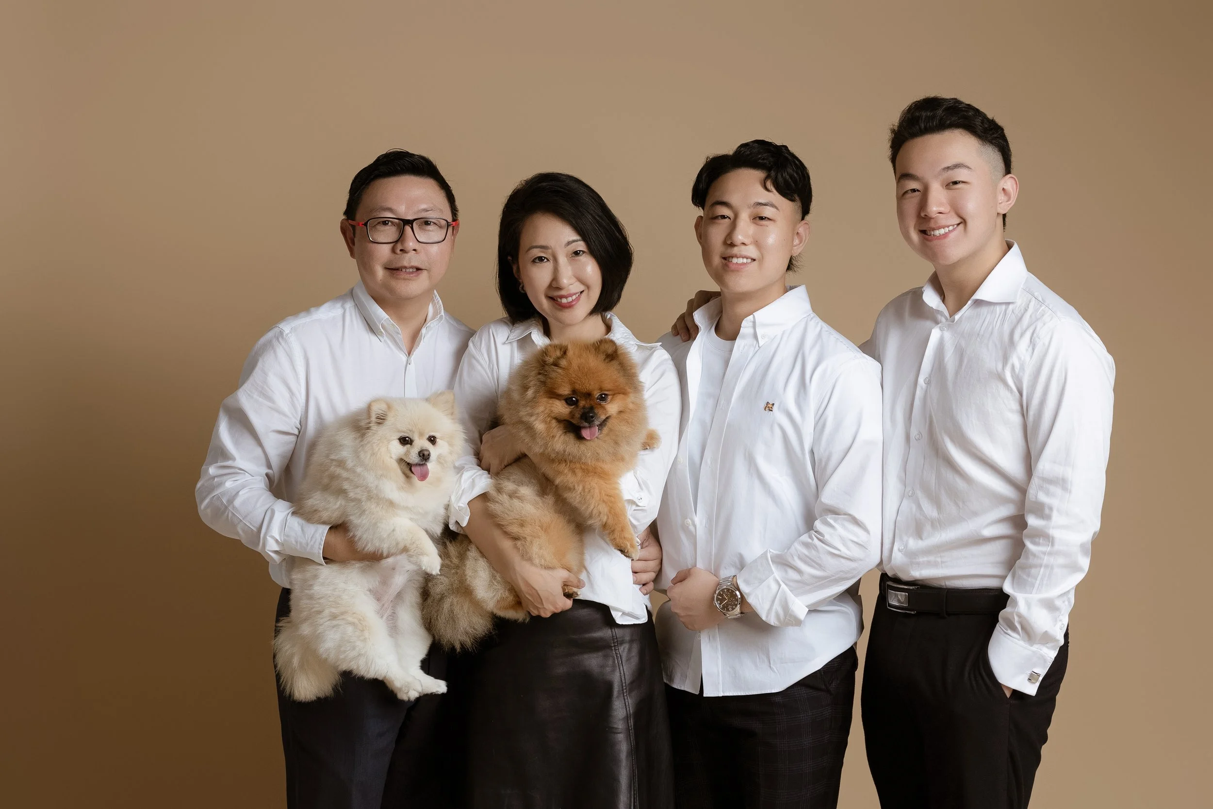 A family of five with two dogs posing together against a beige background. The family members are smiling and dressed in white shirts, with the woman in a leather skirt and the men in dark pants. The dogs are small, fluffy, and light-colored.