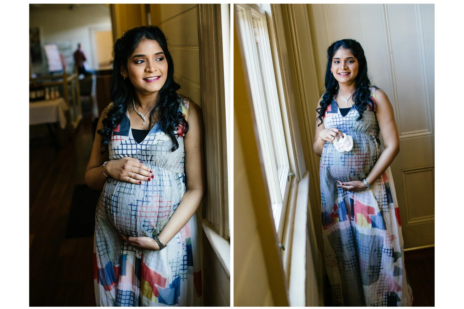A pregnant woman with long black curly hair, wearing a sleeveless, colorful patterned dress, standing indoors near a window, holding her belly with one hand, smiling and looking outside. In the background, there are tables and chairs, and another per