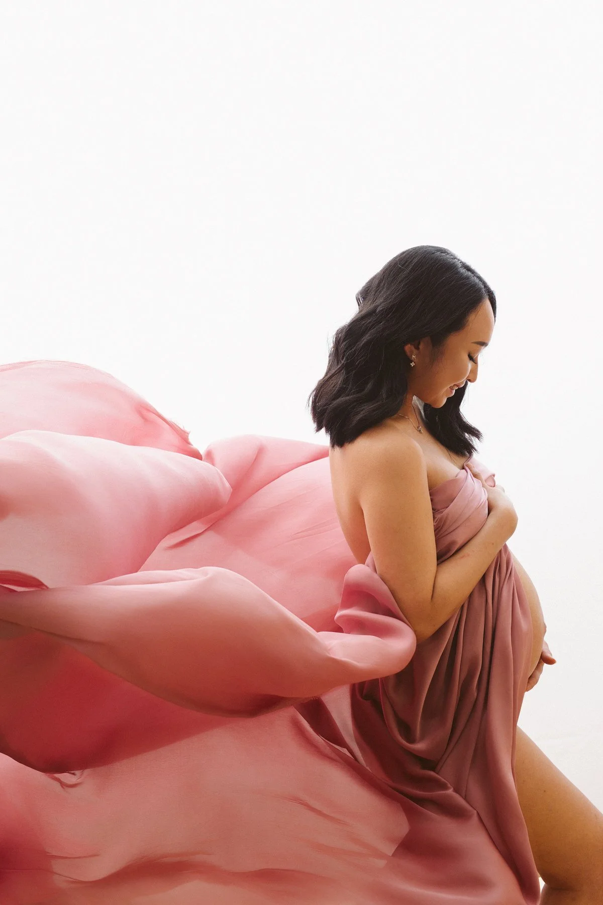 A woman with dark, wavy hair, dressed in a pink gown, poses with her eyes closed and her hand gently placed on her chest, against a white background with large pink fabric resembling a flower.