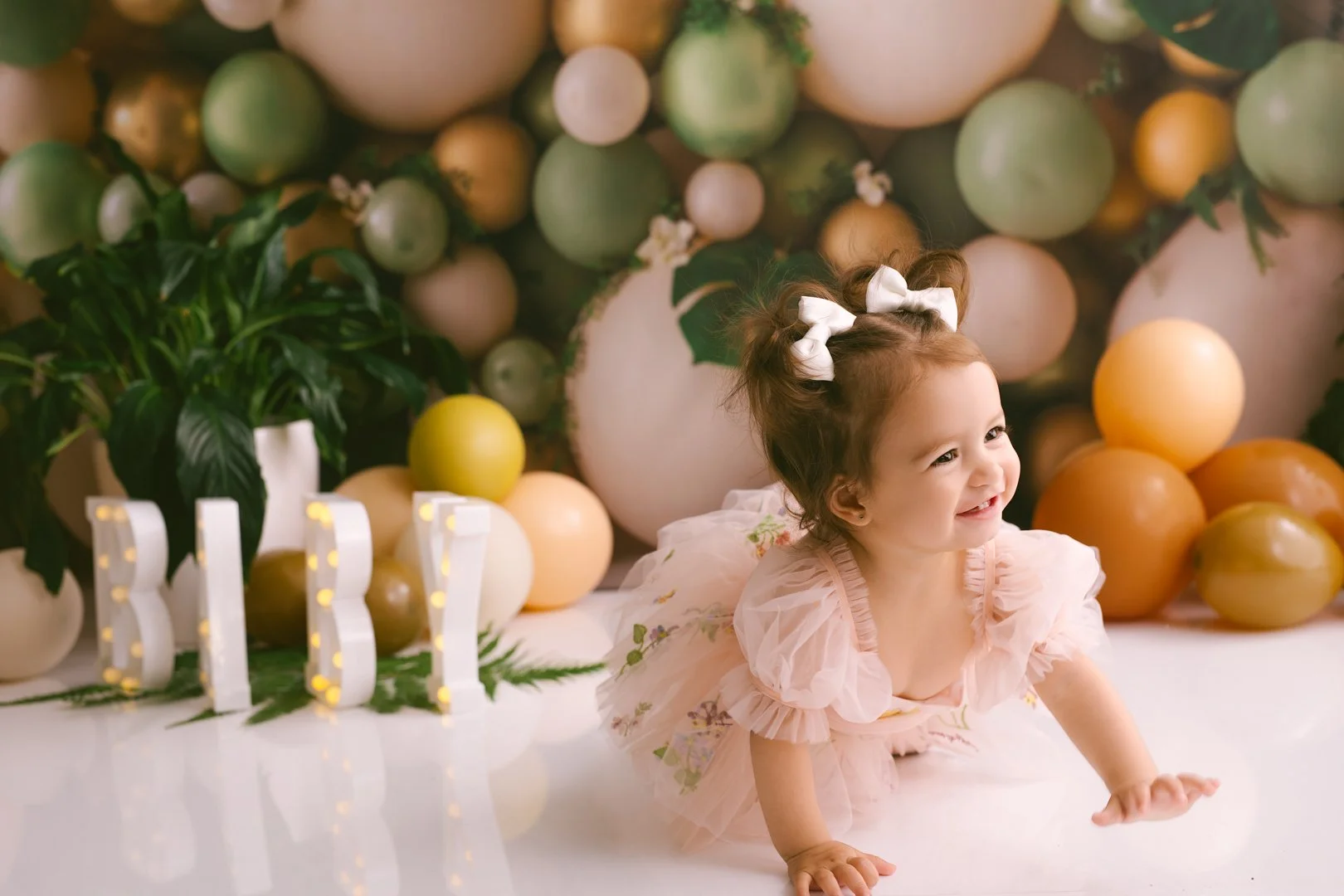 A smiling A  one year old baby girl crawling on the floor during a celebration with pastel-colored balloons and lights spelling 'BABY' in the background.