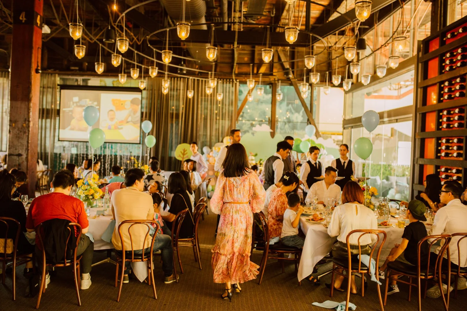 A lively indoor celebration with guests seated at tables, balloons, and festive decorations. A woman in a floral pink dress walks through the room, which features warm lighting, large windows, and a projection screen showing children at a table.