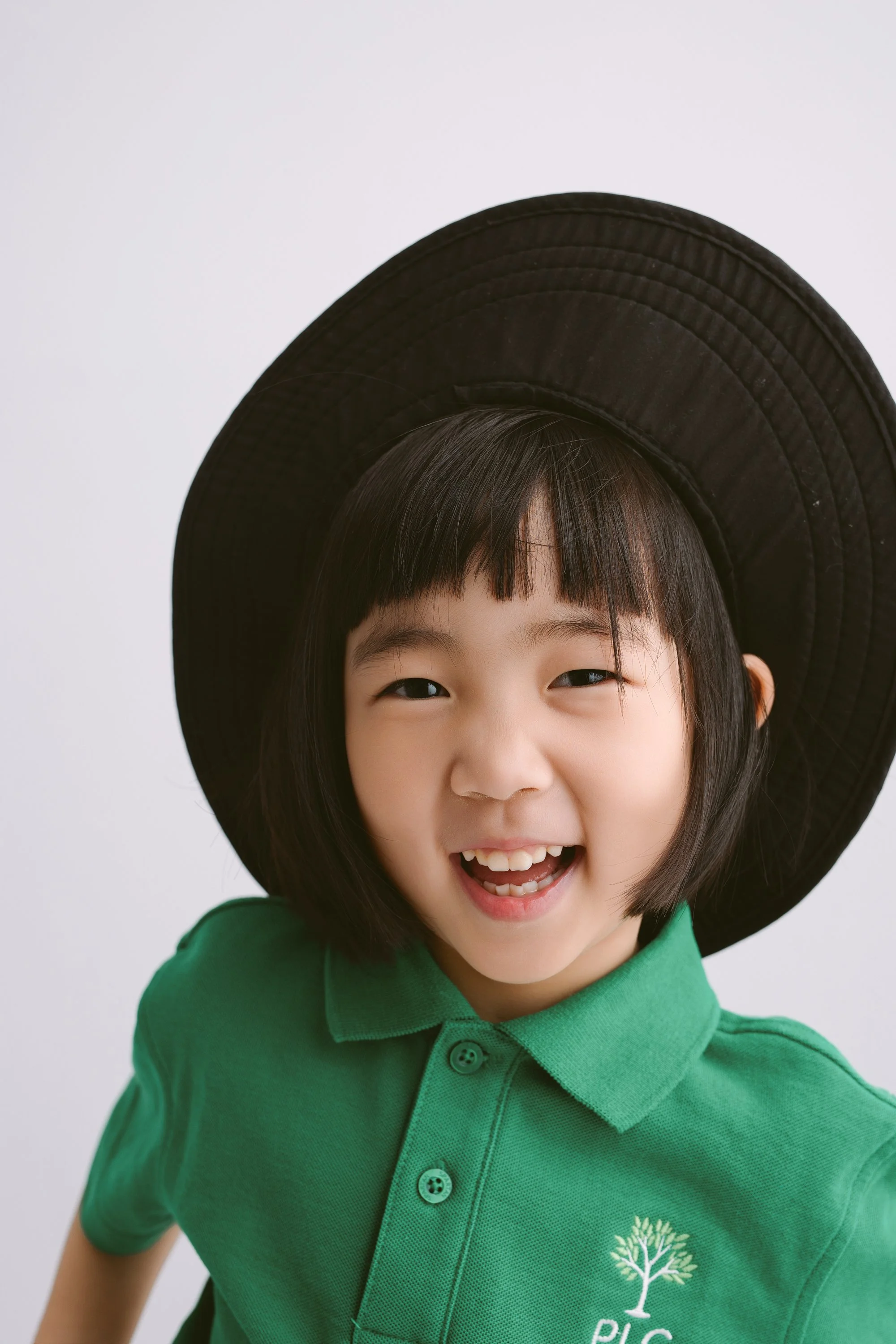 A young girl wearing a large black hat and a green polo shirt with a tree logo and the word 'PIC' smiling and looking at the camera.