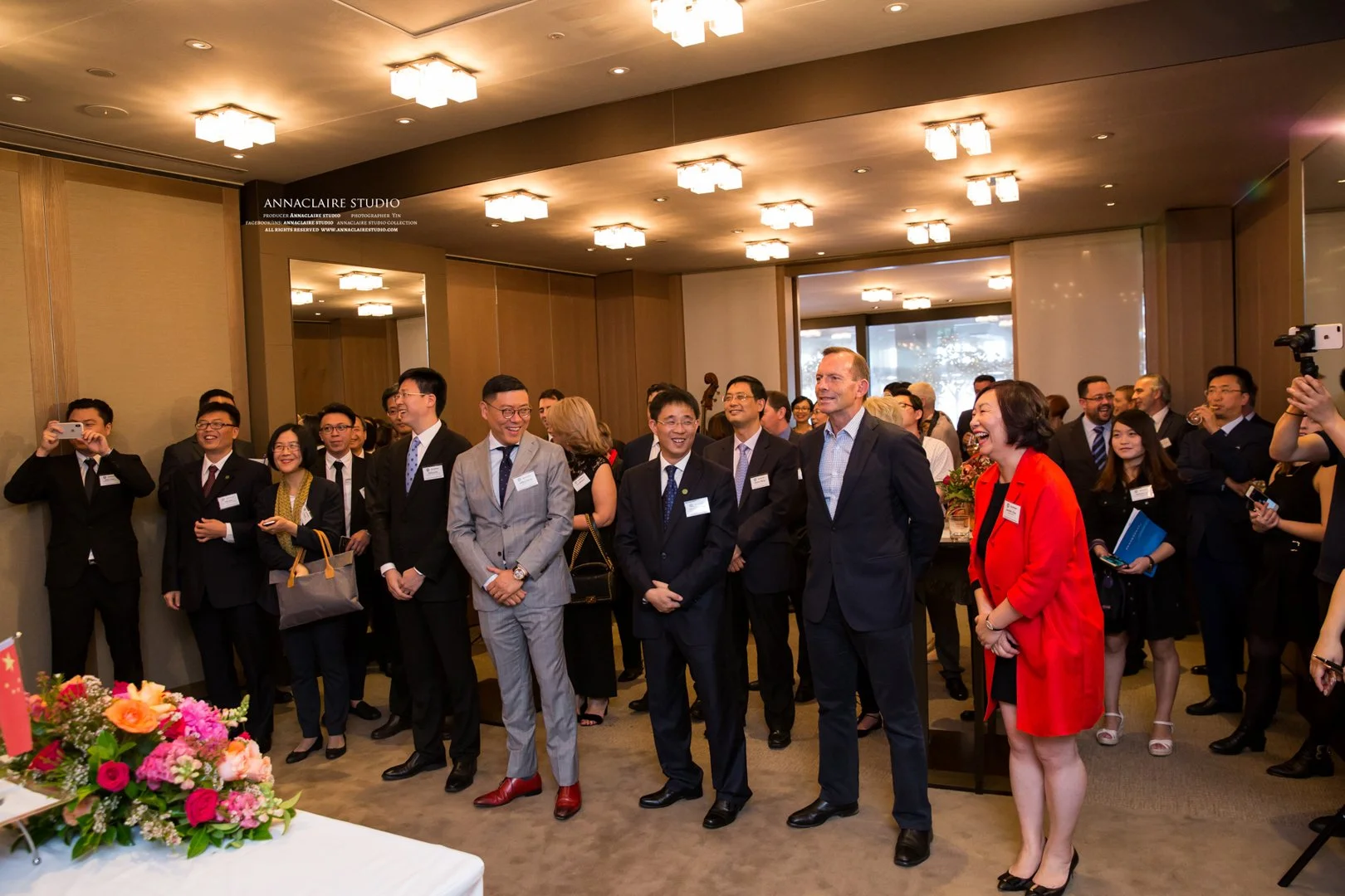 Business professionals gathered at an indoor event, smiling and taking photos, with a floral arrangement on a table in the foreground.