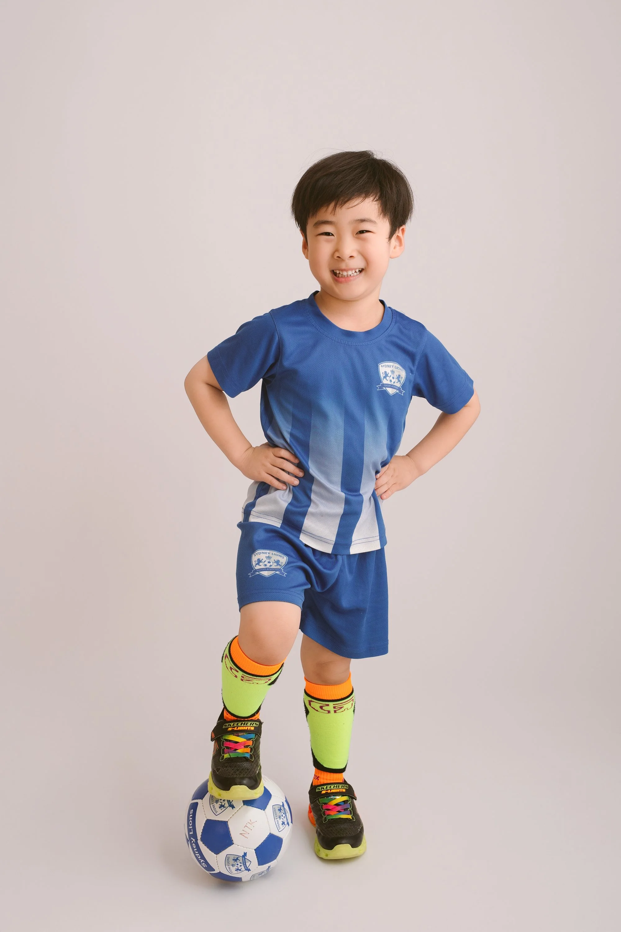 Young boy in blue soccer uniform standing with hands on hips, smiling, with one foot on a soccer ball against a plain white background.