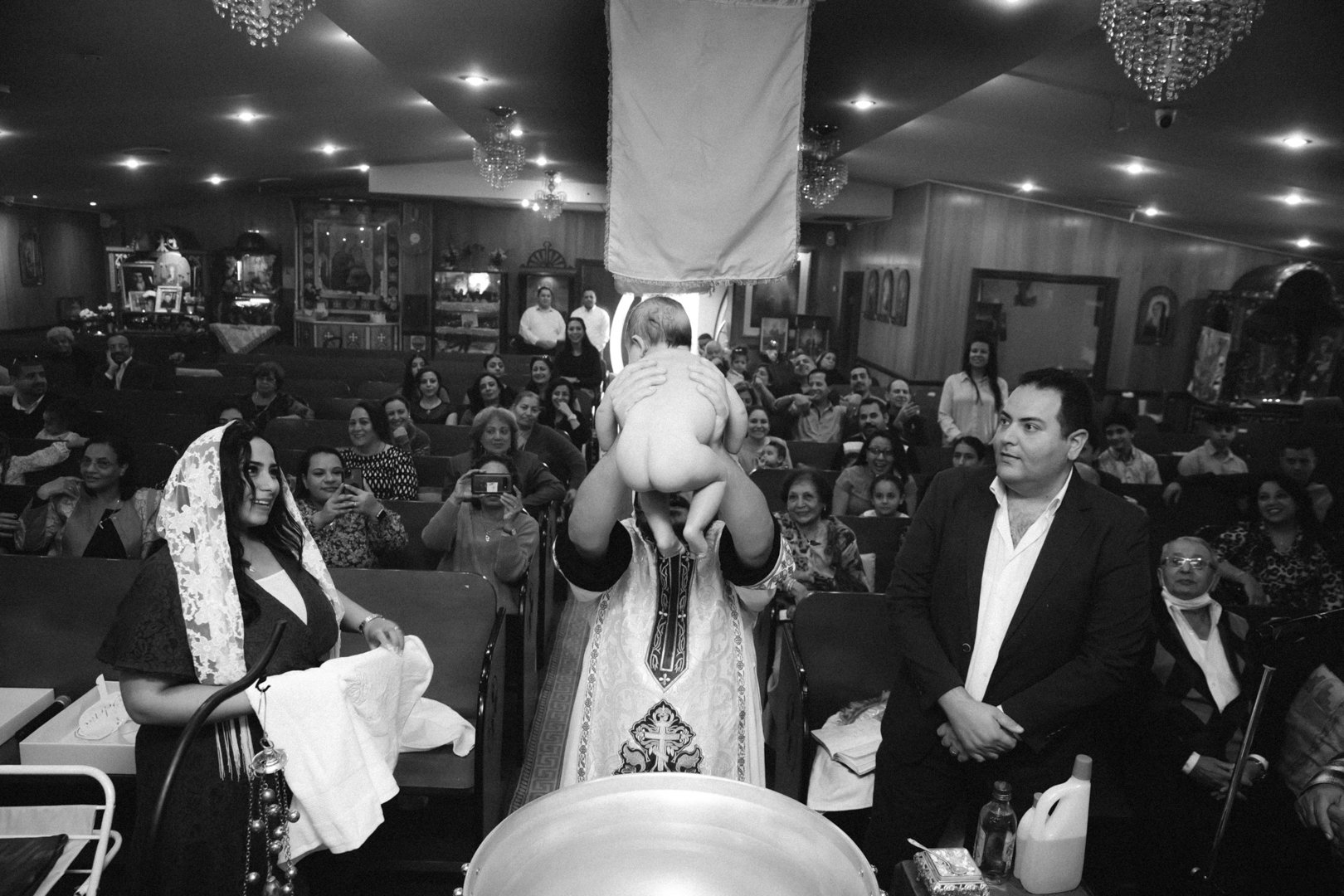 A religious ceremony inside a church with a priest or clergyman holding a baby in front of an audience. The audience is watching the event, some taking photos. The room is decorated with chandeliers and religious icons on the walls.