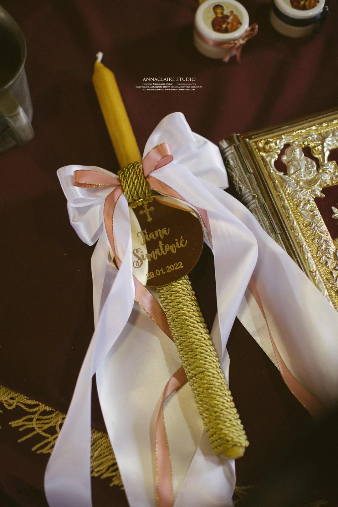 A decorated candle with a white and pink ribbon bow, a gold inscription tag with a cross that reads 'Tanja Simatović 29.01.2022,' and some religious icons and a golden ornate frame on a table.
