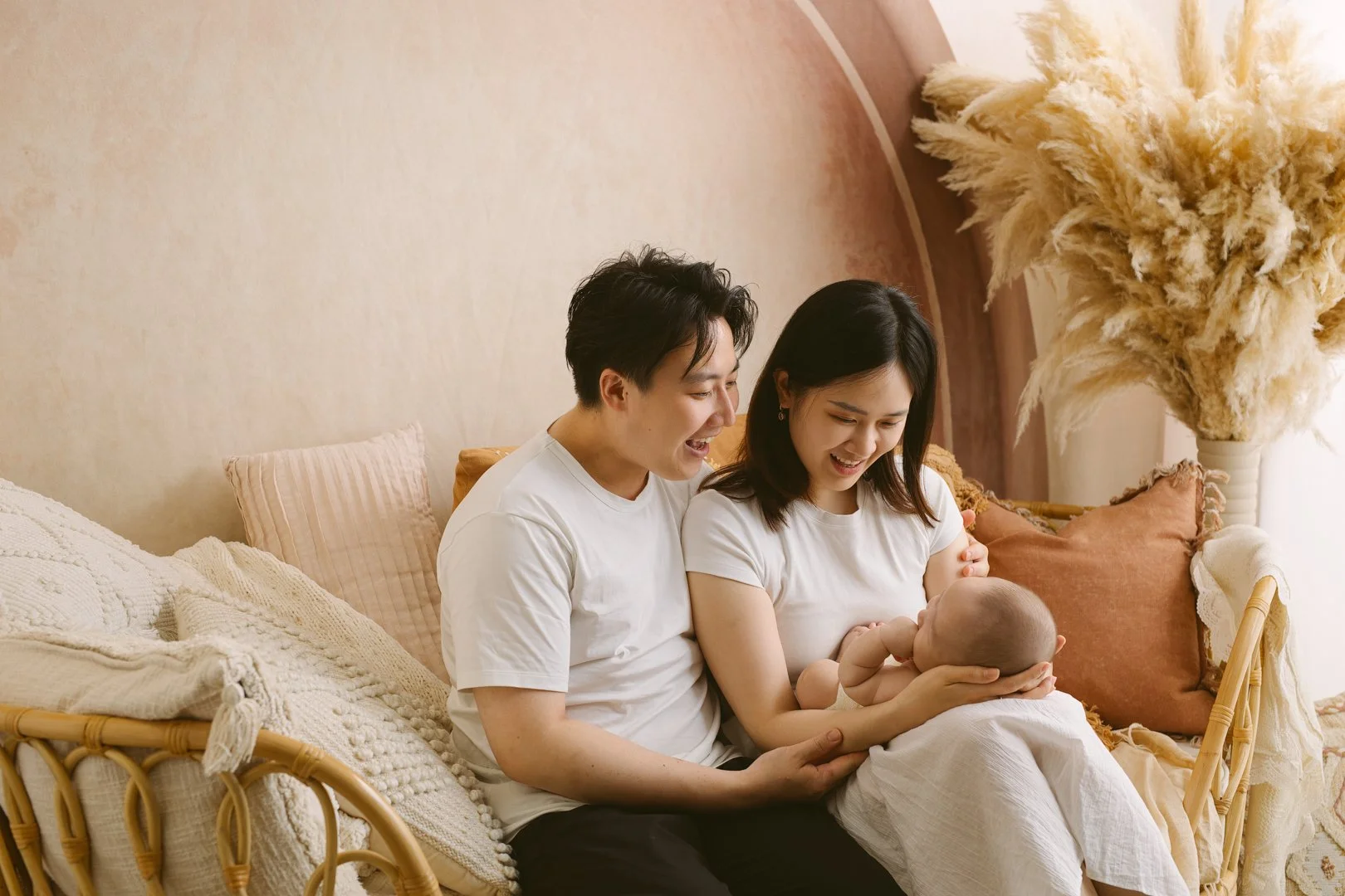 A family sitting on a beige couch, consisting of a man, woman, and baby. The man and woman are smiling and looking at the baby, who the woman is holding and breastfeeding. The room has soft lighting, and decorative pampas grass is visible in the back