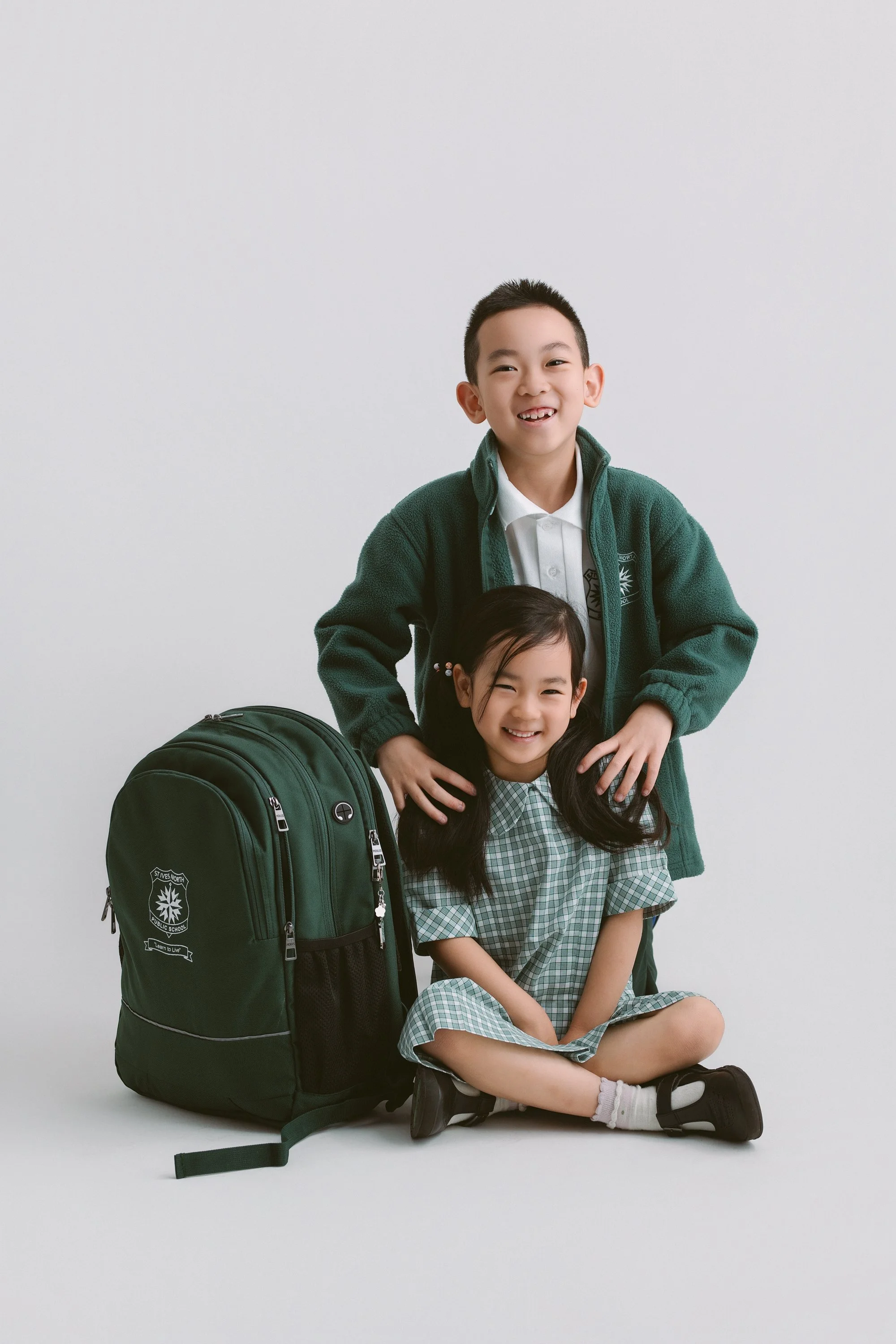 A boy and a girl in school uniforms smiling, with the boy standing behind and the girl sitting on the floor cross-legged, next to a green backpack with a school emblem against a plain white background.