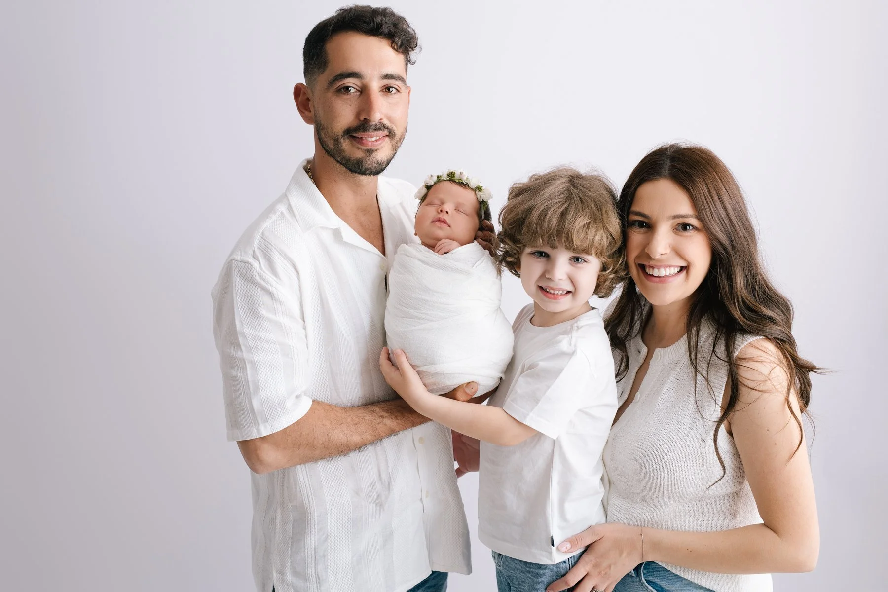 Family portrait of a man, woman, a young boy, and a newborn baby girl, with the baby wrapped in white cloth and wearing a floral headband, standing against a plain light gray background.