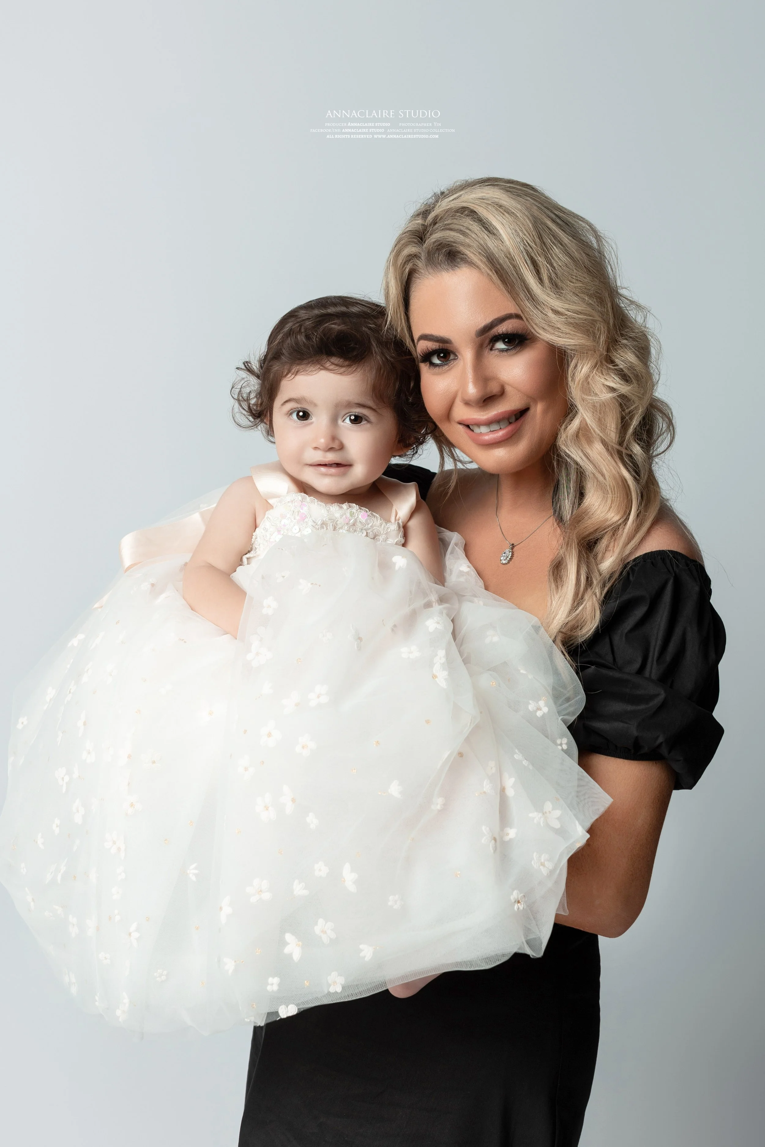 A woman holding a young girl dressed in a white dress with floral details. Both are smiling toward the camera against a plain background.