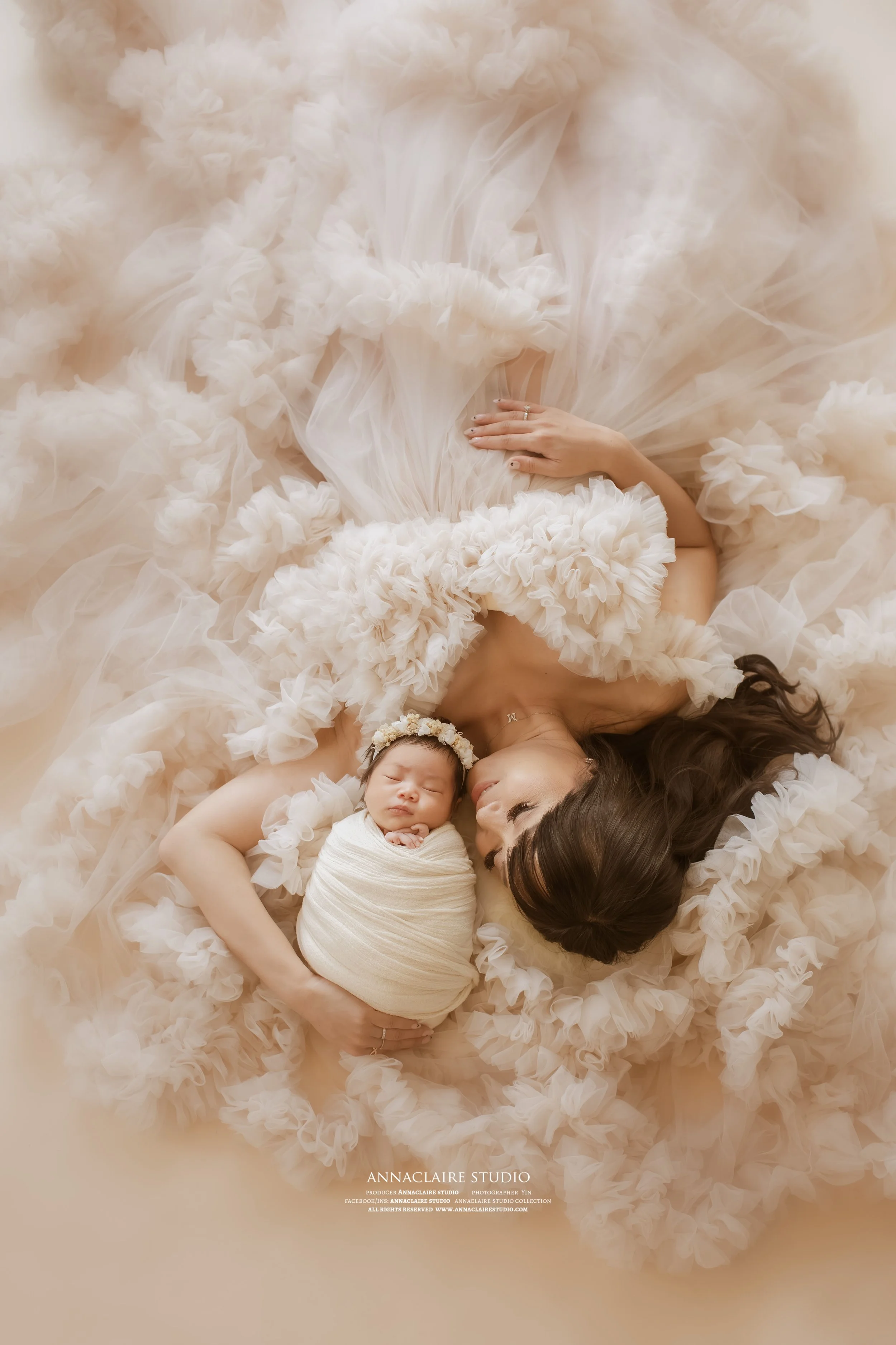 A new mum  and a newborn baby are lying down on a fluffy, ruffled fabric. The woman is gently holding the baby, who is wrapped in a white cloth and wearing a flower headband. They are facing each other, with the woman gazing at the baby.