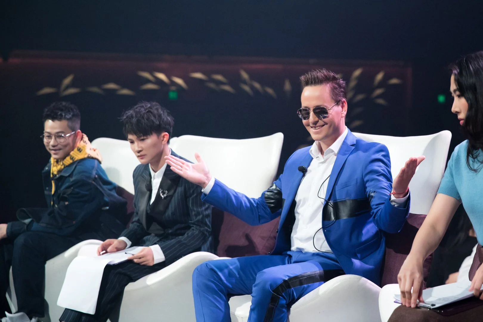 Four people sitting on stage during a panel discussion or event, with the man in the blue suit smiling and gesturing with his hands.