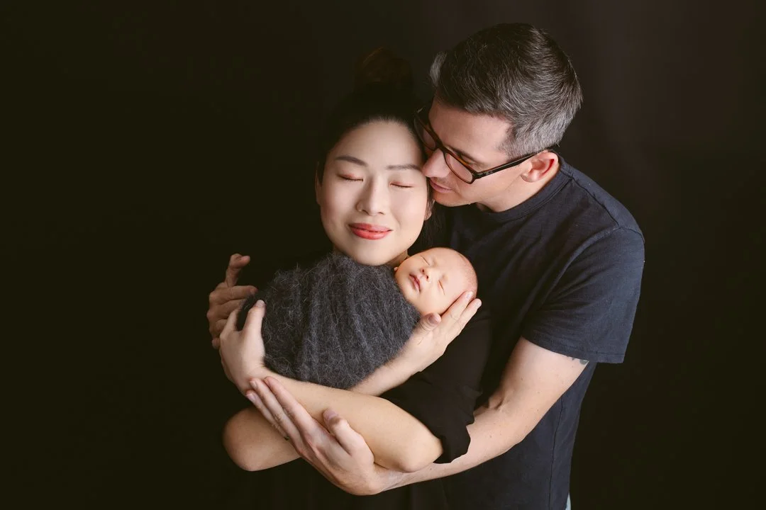 A family portrait of a woman, man, and a newborn baby against a black background. The woman is holding the baby close, and the man is leaning in to kiss the woman's temple, all with happy expressions.