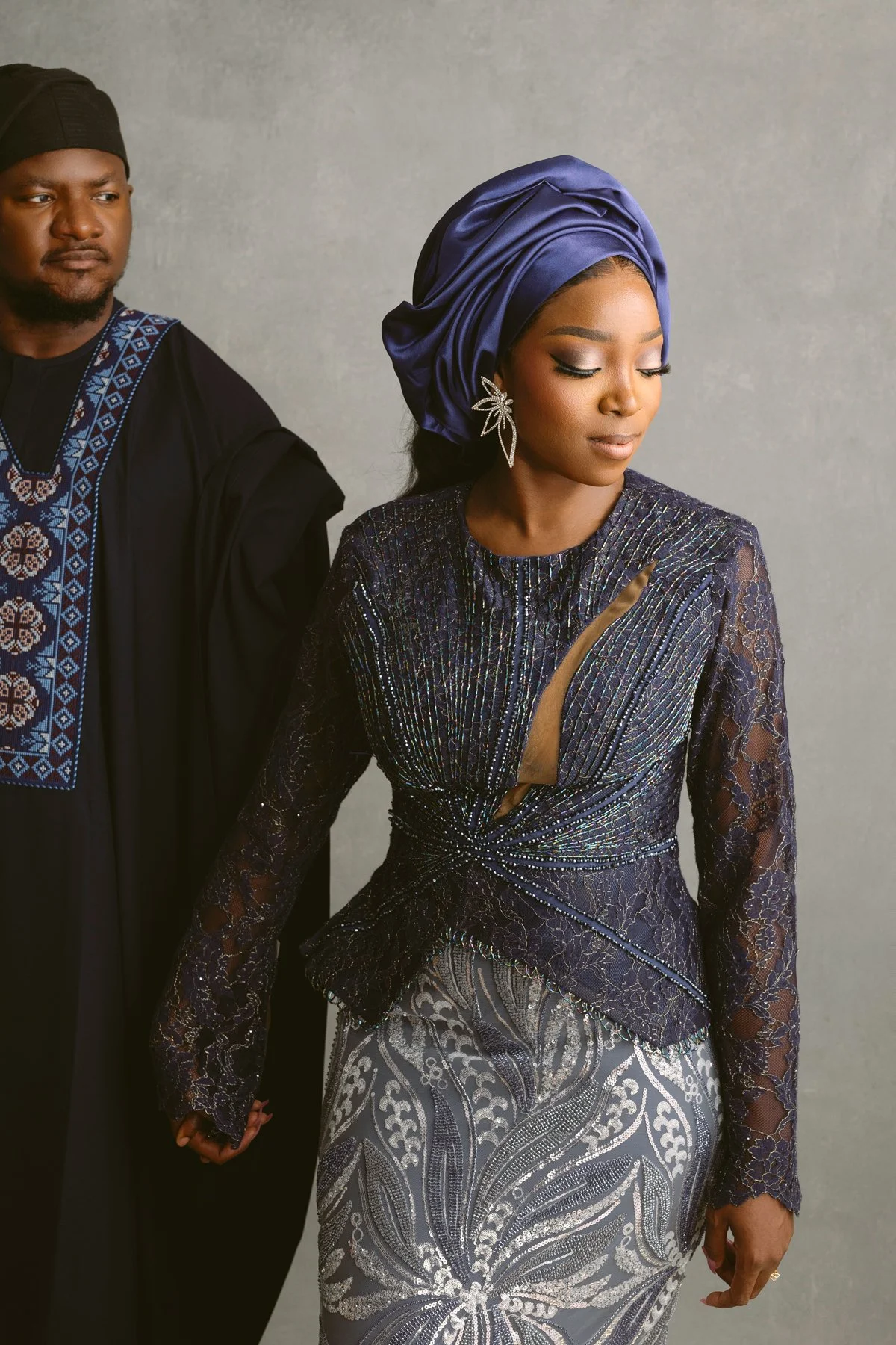A woman dressed in a dark, embellished dress with a slit and lace sleeves, wearing a blue head wrap and large floral earrings, holding hands with another person dressed in traditional African attire with embroidery.