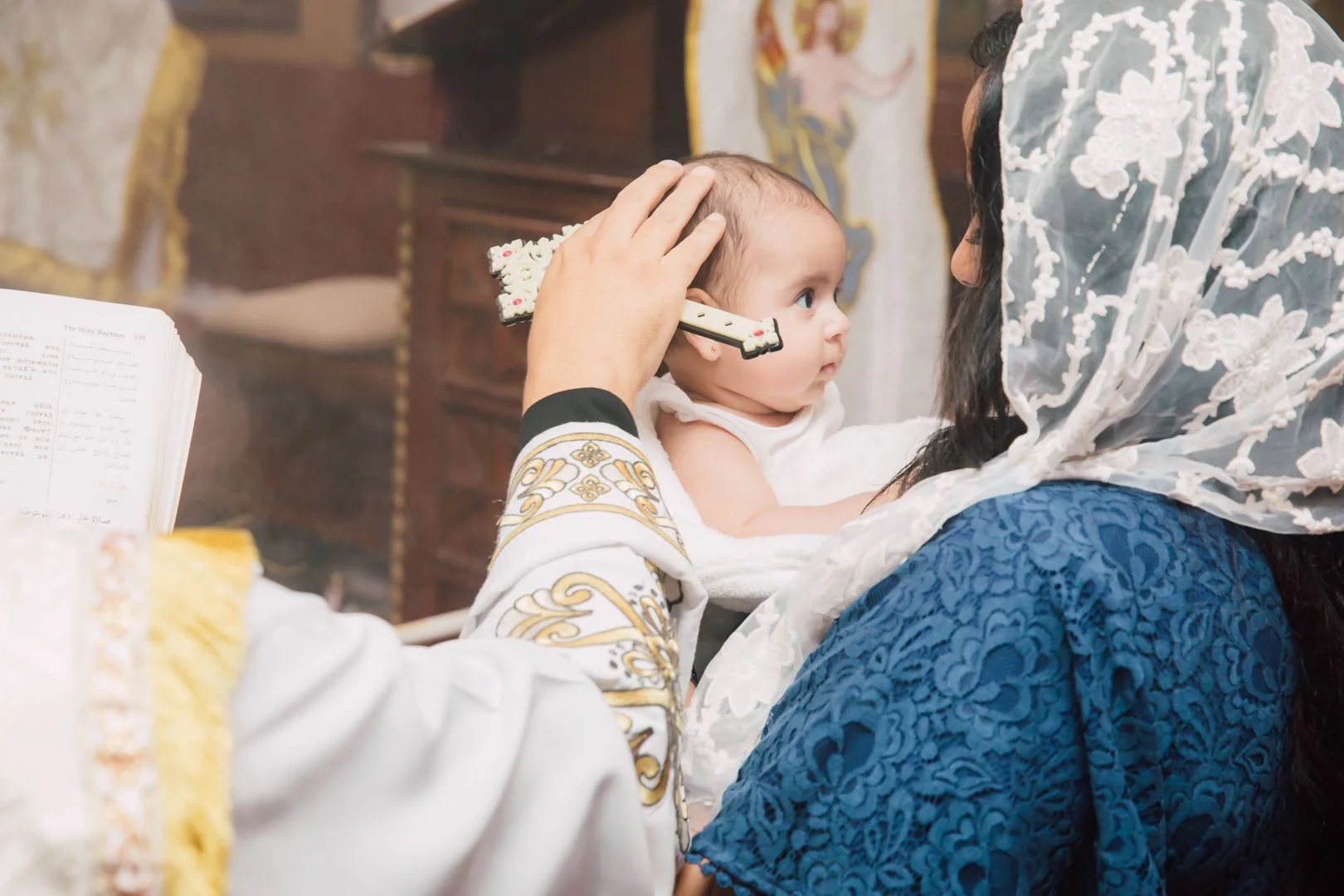 A baby being blessed during a religious ceremony, held by a woman wearing a lace head covering.