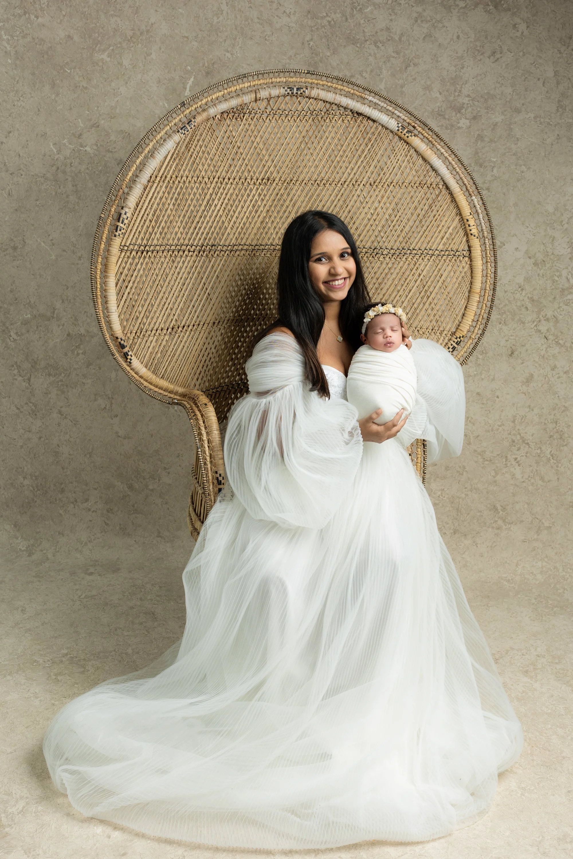 a new mum  in a white dress holding a sleeping baby wrapped in a white blanket, seated on a wicker chair with a large woven backrest, in a studio setting.