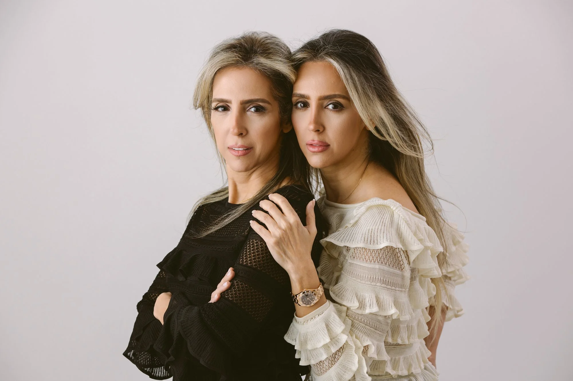 Two women with long hair standing close together against a plain background, one in black and the other in white, with confident expressions.