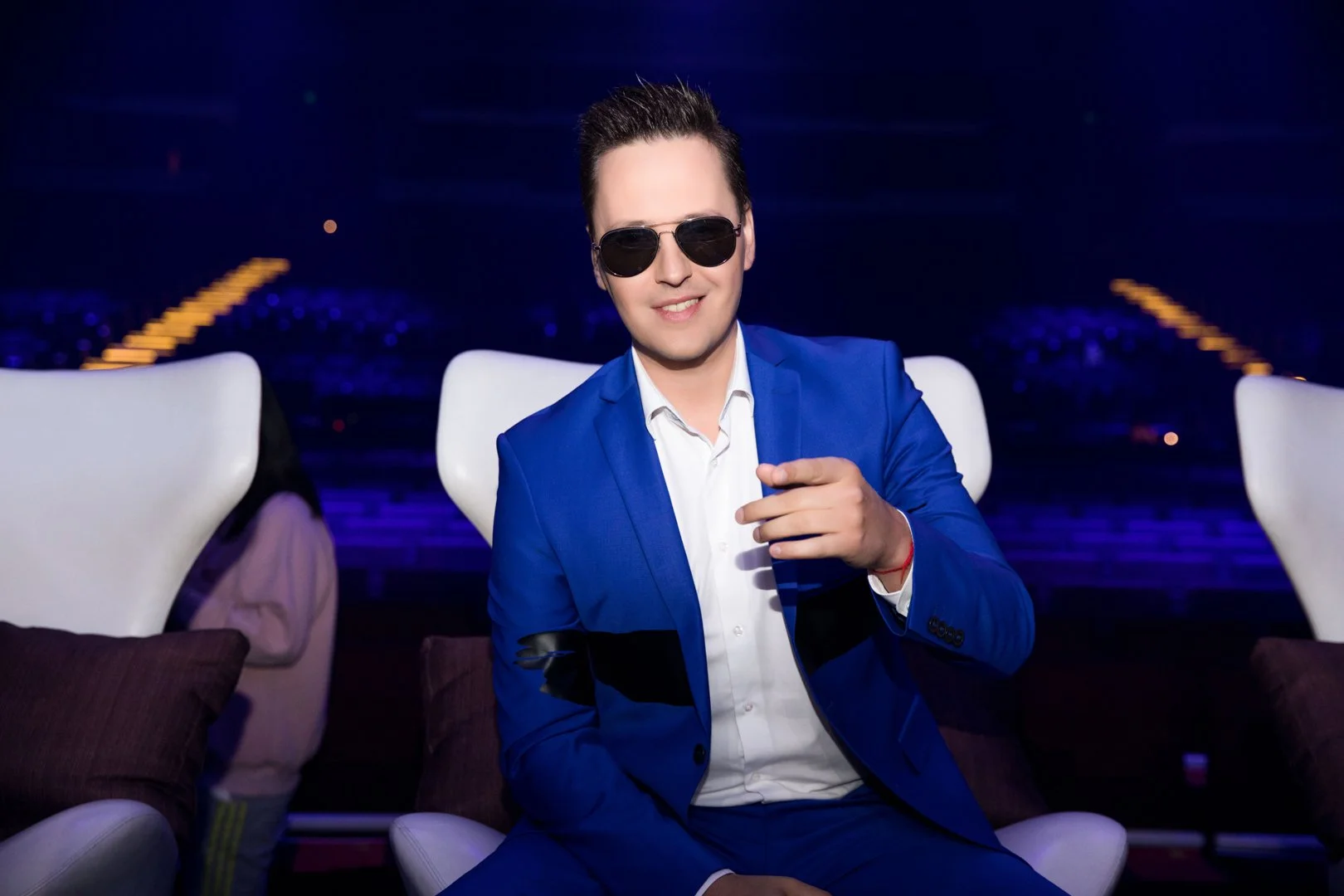 A man in a blue suit and sunglasses sitting on a white chair, pointing at the camera, with a dark background and illuminated stairs.