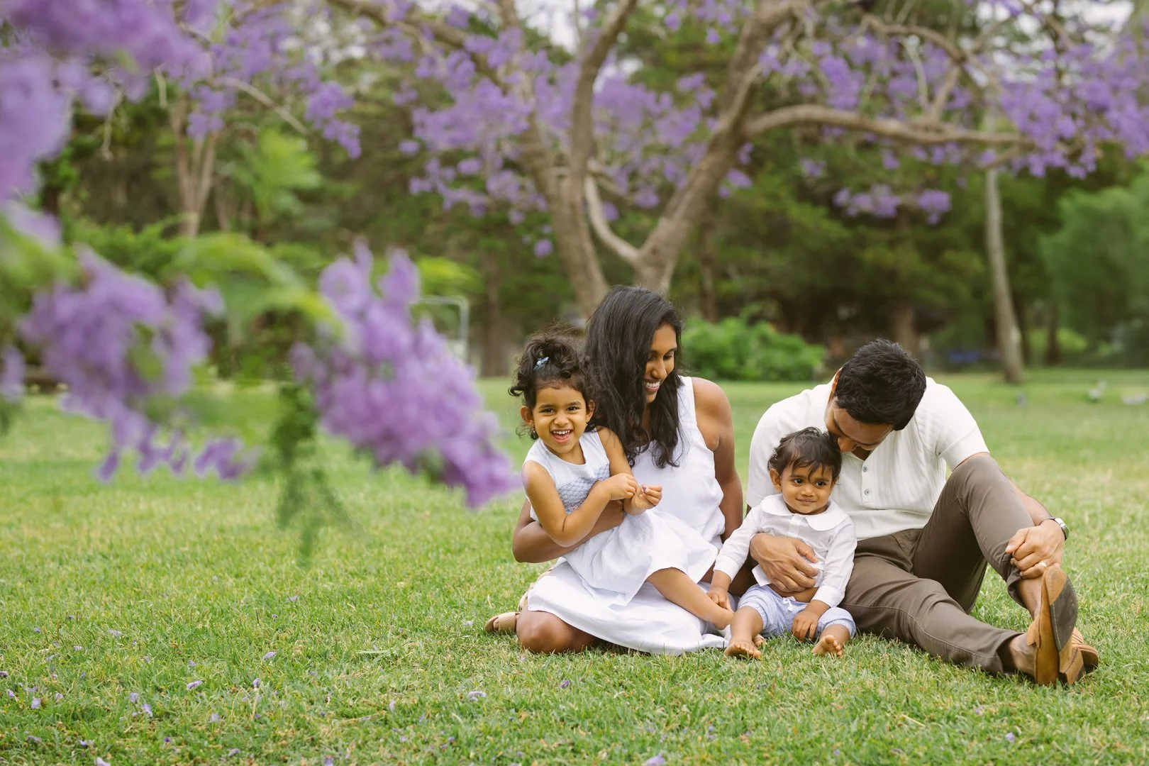 A family of four sitting on the grass in a park with purple flowering trees in the background. The mother and father are with two young children, one girl and one boy, all smiling and enjoying the outdoors.