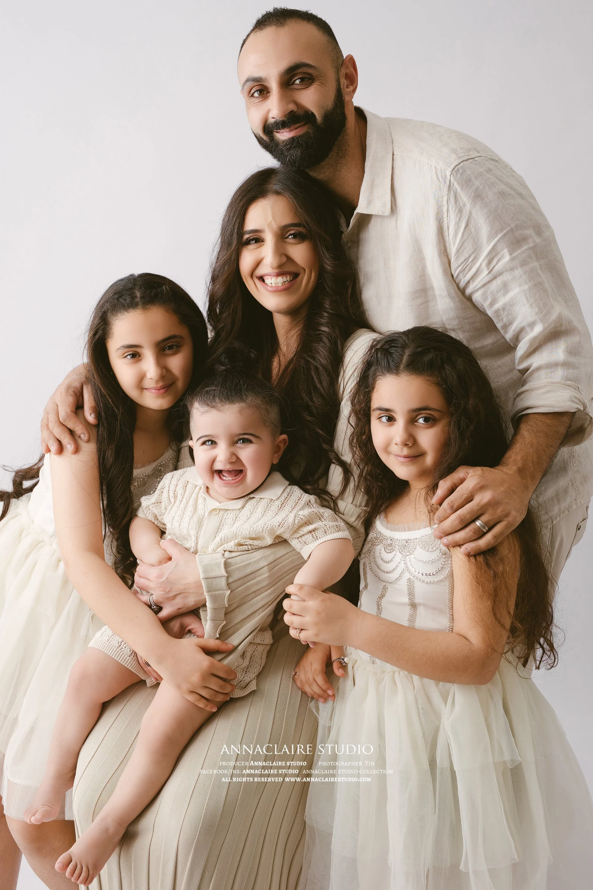 A happy family of five, including mum , dad, 2 sisters,and a cute baby brother  smiling and posing together in a studio with a white background.
