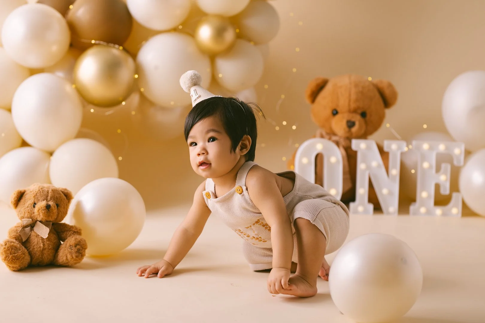 1 yr old baby boy  crawling on the floor in a birthday celebration setting with white and gold balloons, teddy bears, and illuminated letter signs spelling 'ONE' in the cake smash photo background.