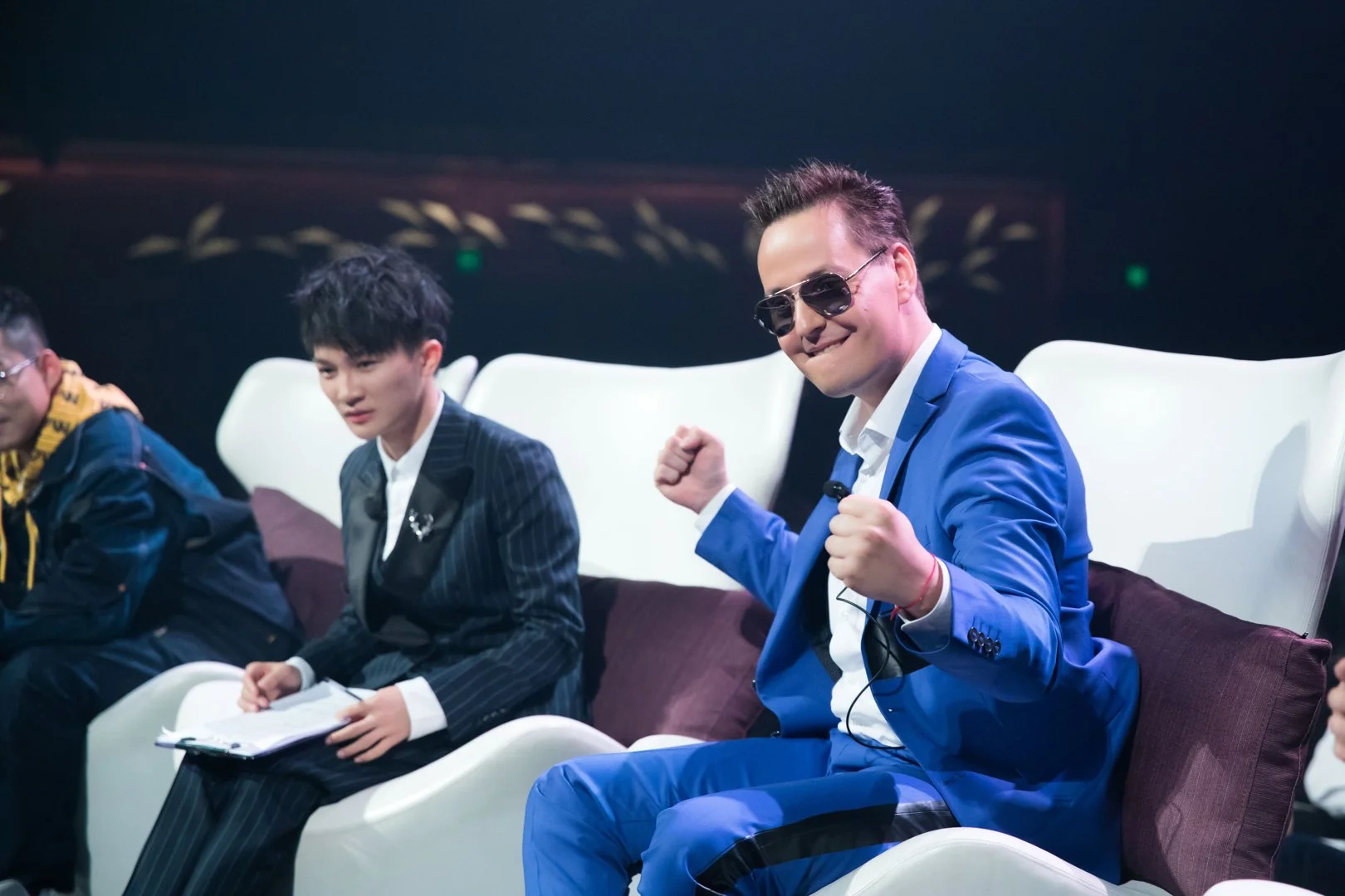 A group of people sitting on white chairs in a dark room, with one man in a blue suit and sunglasses smiling and making a fist gesture.