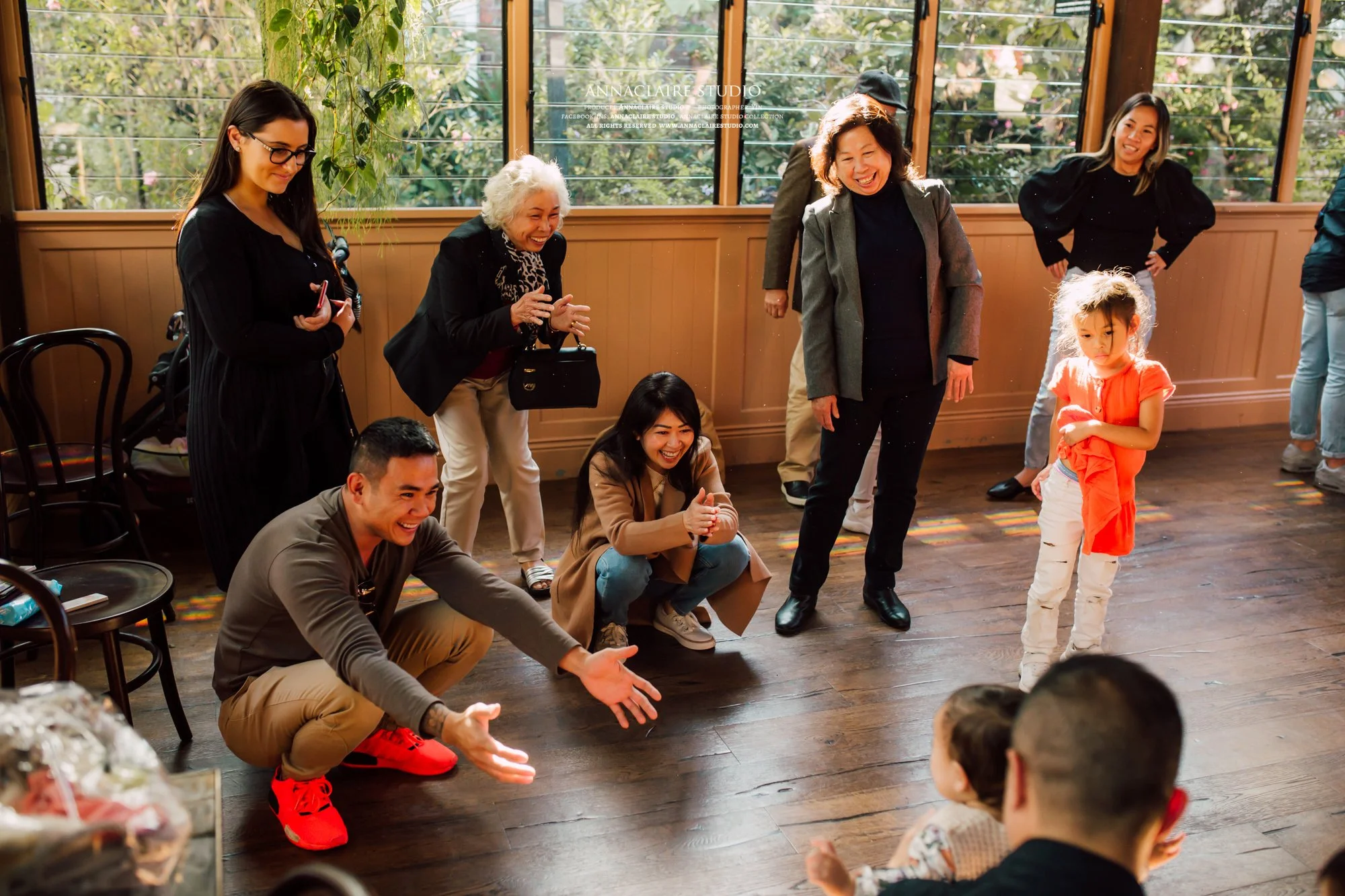 A group of adults and children are gathered indoors, with some standing and others squatting, engaging in a playful activity. They are smiling and appear to be enjoying themselves. The room has large windows with natural light, wooden flooring, and s