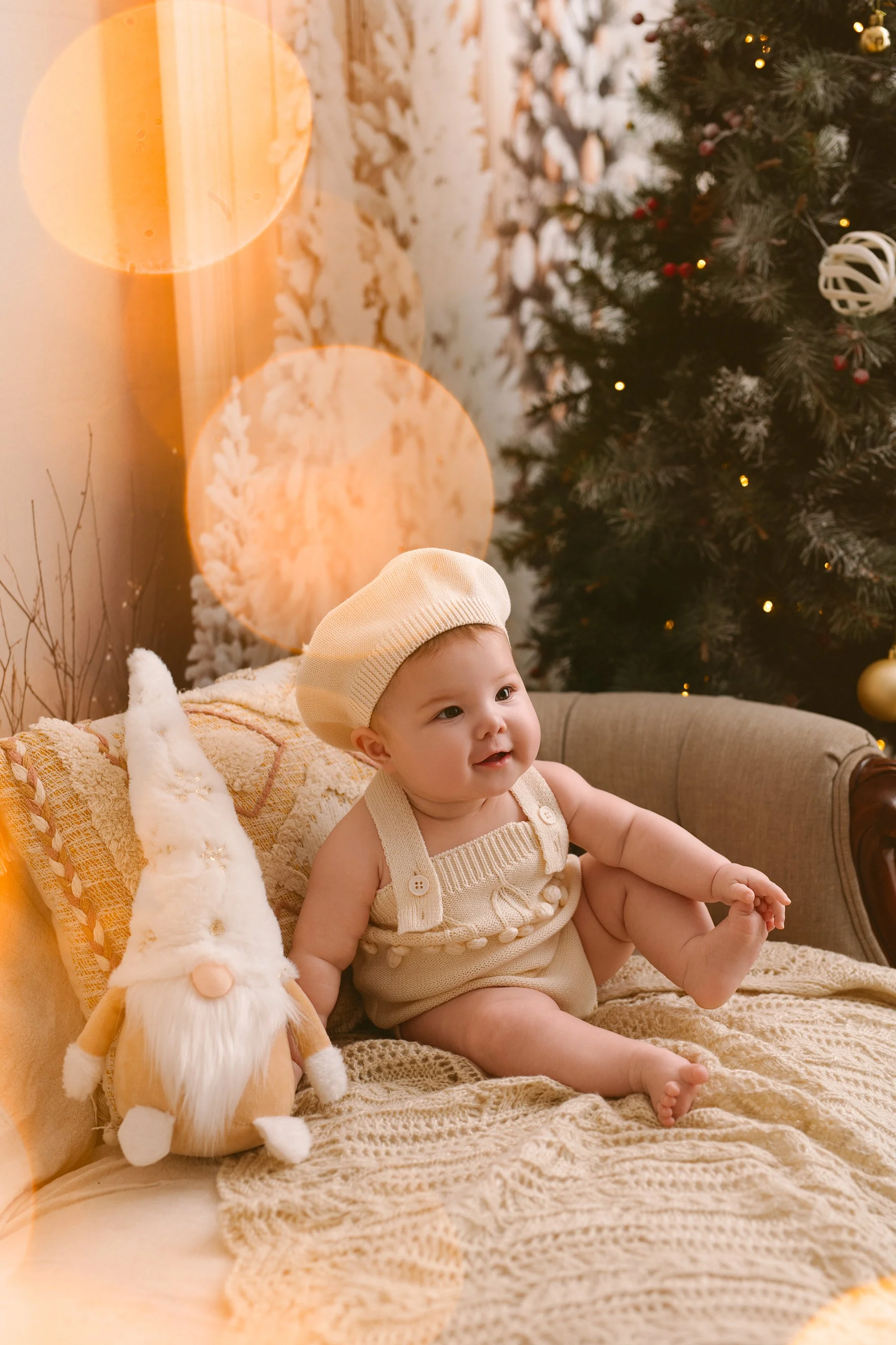 A baby sitting on a couch next to a Christmas tree decorated with ornaments, with a stuffed gnome toy and a cozy throw blanket.