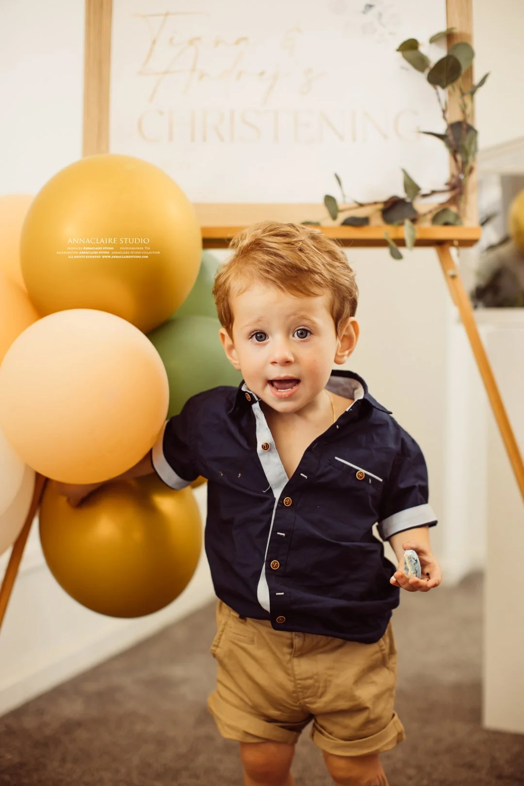 A young boy with red hair and blue eyes is standing in front of a balloon decoration in a room. He is wearing a navy blue shirt with rolled-up sleeves and tan shorts. The background includes a sign that reads 'Christening' and some greenery.