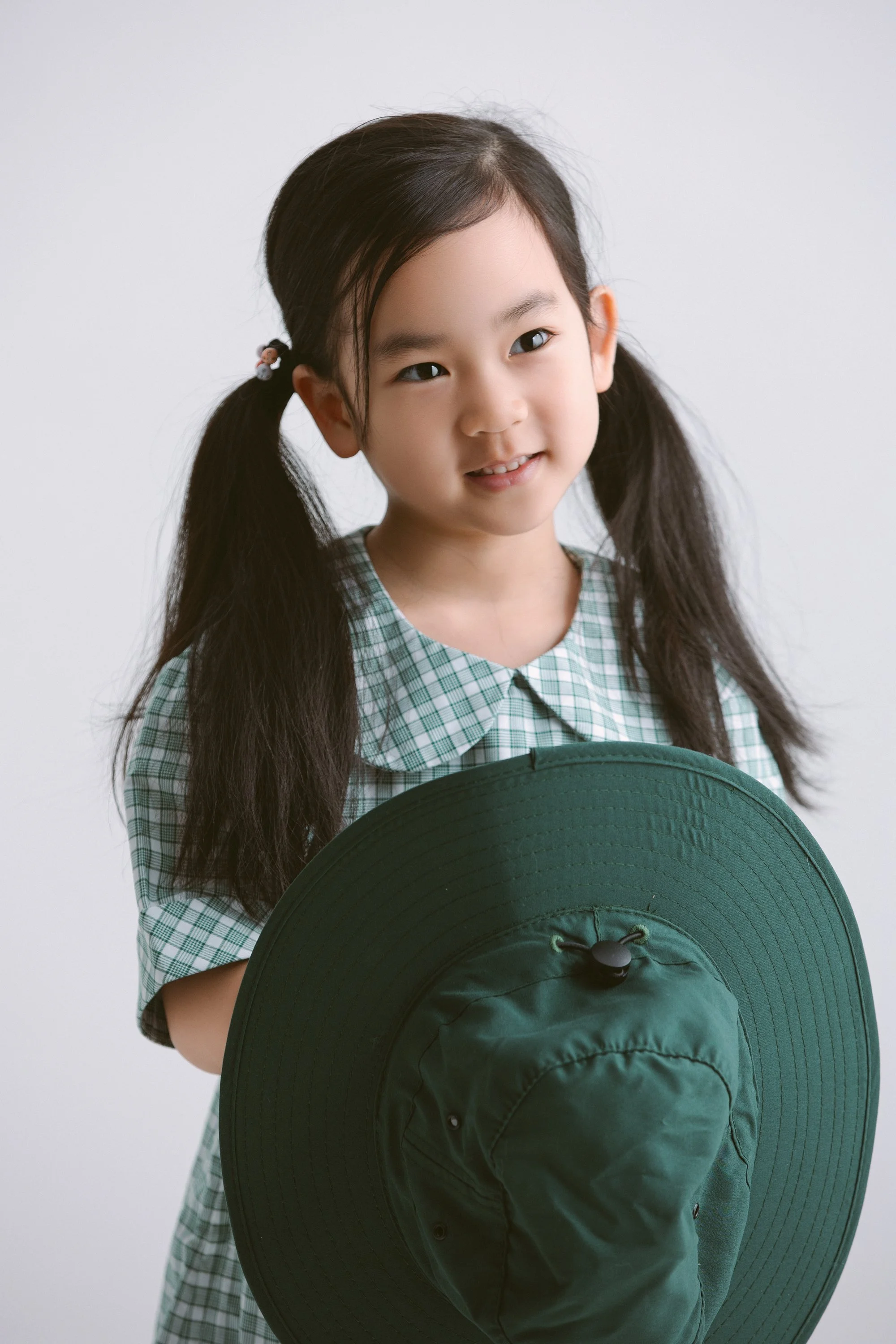A young girl with long dark hair in pigtails, wearing a green and white checkered shirt, holding a dark green wide-brimmed hat, looking at the camera with a slight smile.