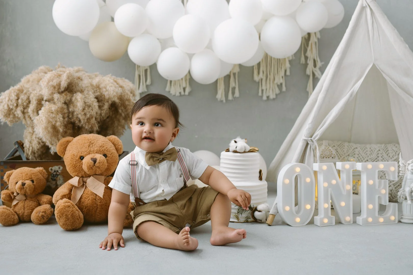 1 year old baby Baby boy sitting on the floor with toys and decorations, including a white tent, teddy bears, cotton plants, and illuminated "ONE" sign, in a neutral-colored room.