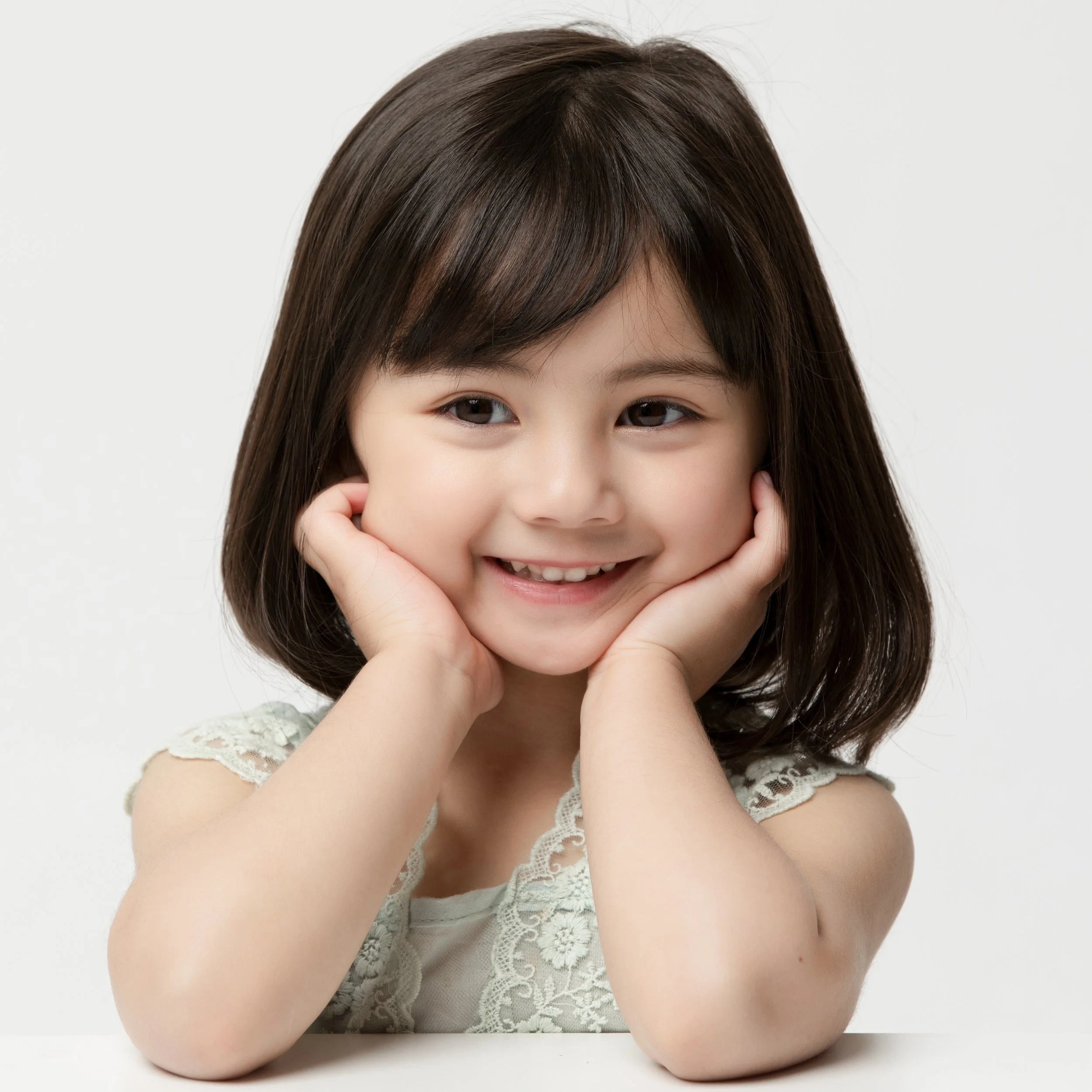 A young girl with shoulder-length dark hair, smiling with her hands on her cheeks, wearing a white lace top, against a plain light background.