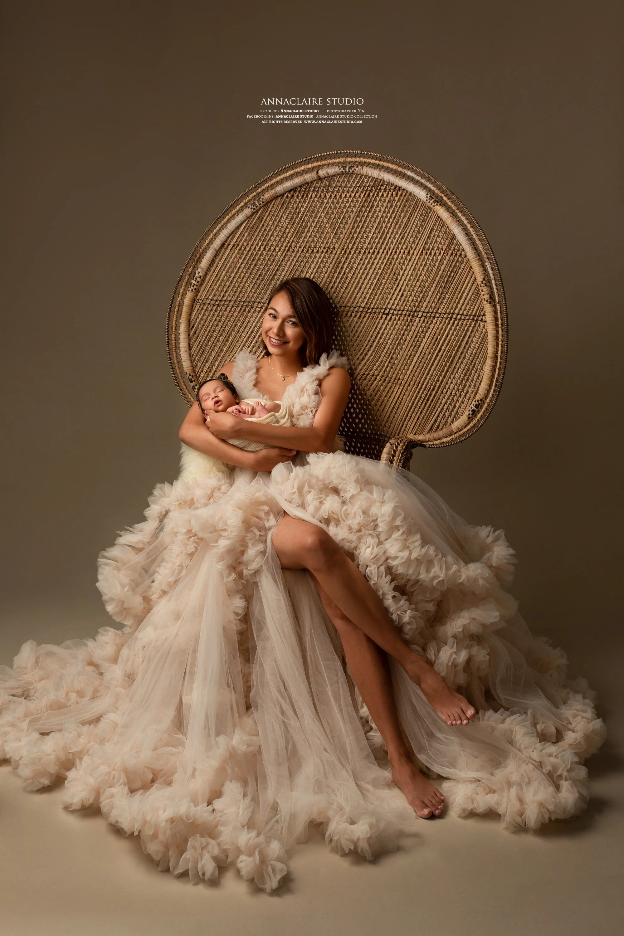 the new mum dressed in an elaborate, flowing cream-colored gown with ruffled layers, sitting on the floor and holding a sleeping baby with a floral headband while sitting on a wicker chair against a plain background.