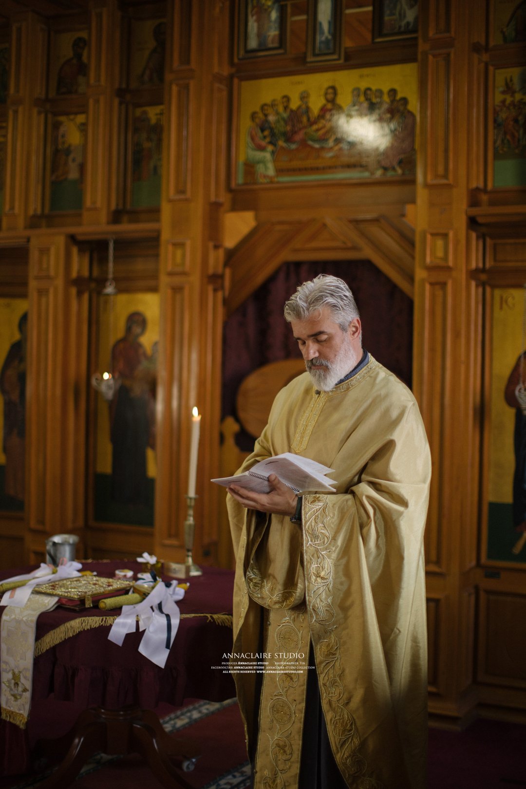A priest in gold vestments reading from a book during a religious service inside a church with wooden icons and paintings on the walls.